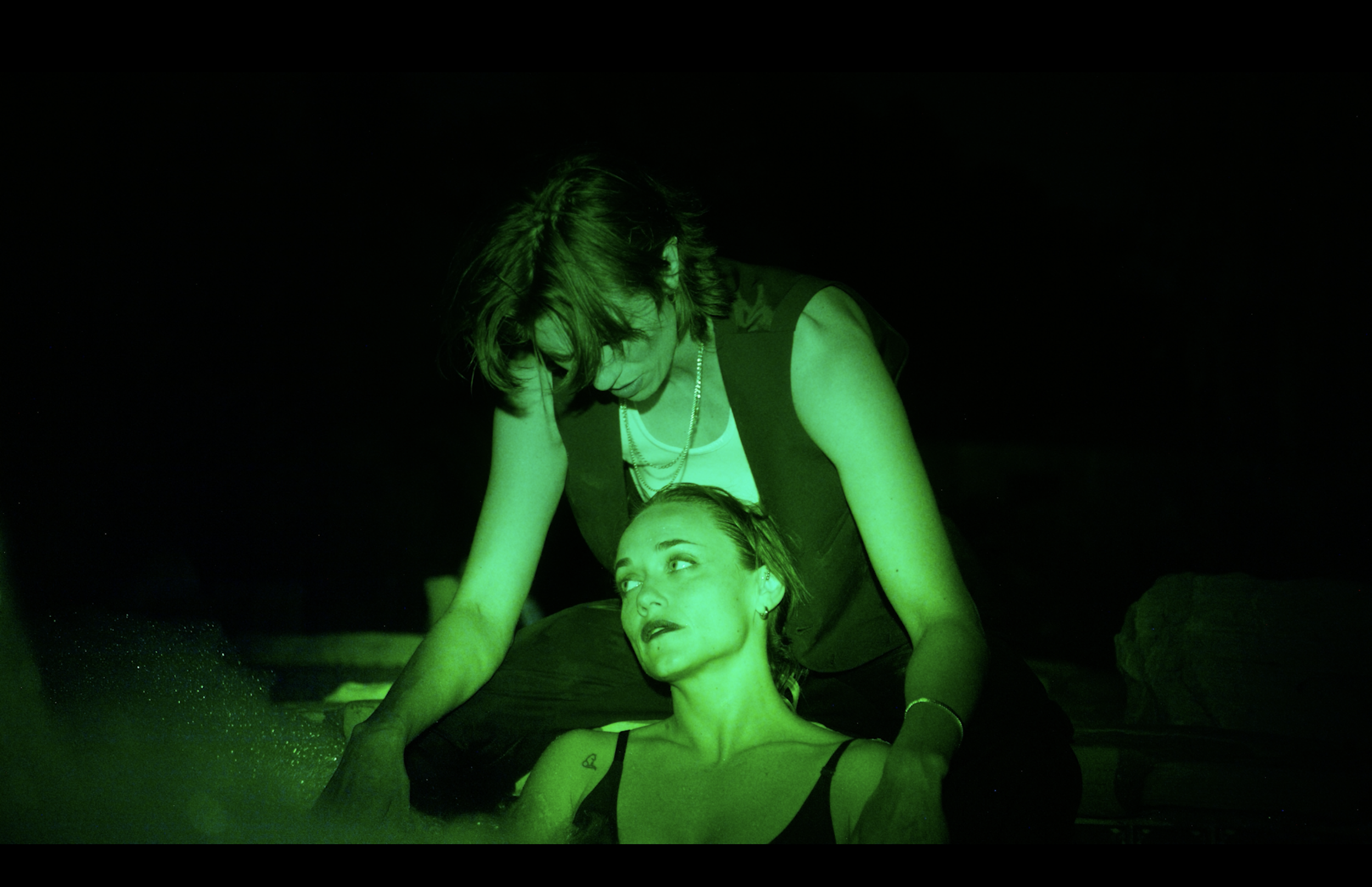Two women in a dark, green-lit environment, with one woman lying down and the other woman leaning over her, appearing to support or assist her.