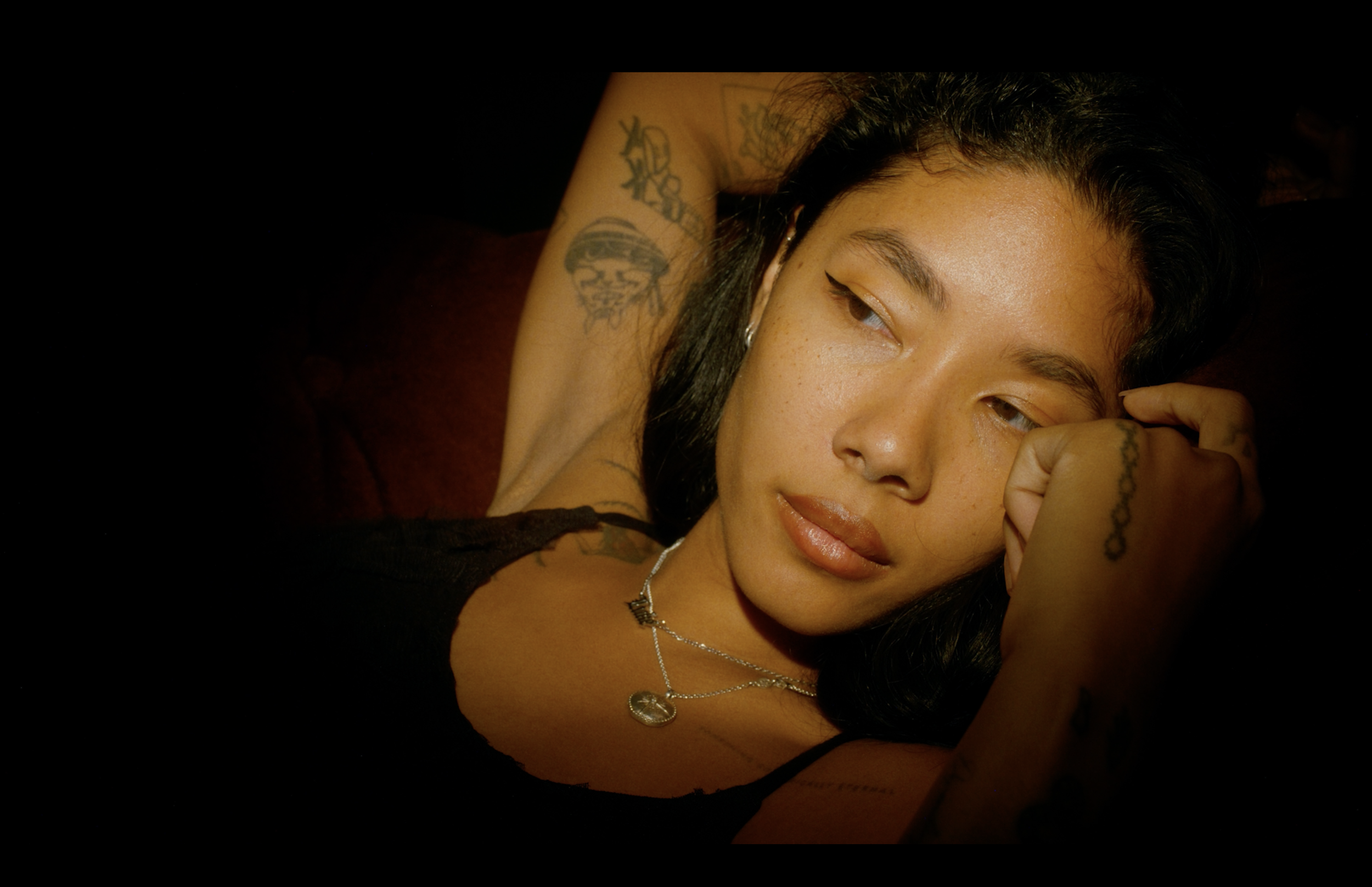 A young woman with dark hair and tattoos resting her head on her arm, looking away from the camera in a dimly lit setting.