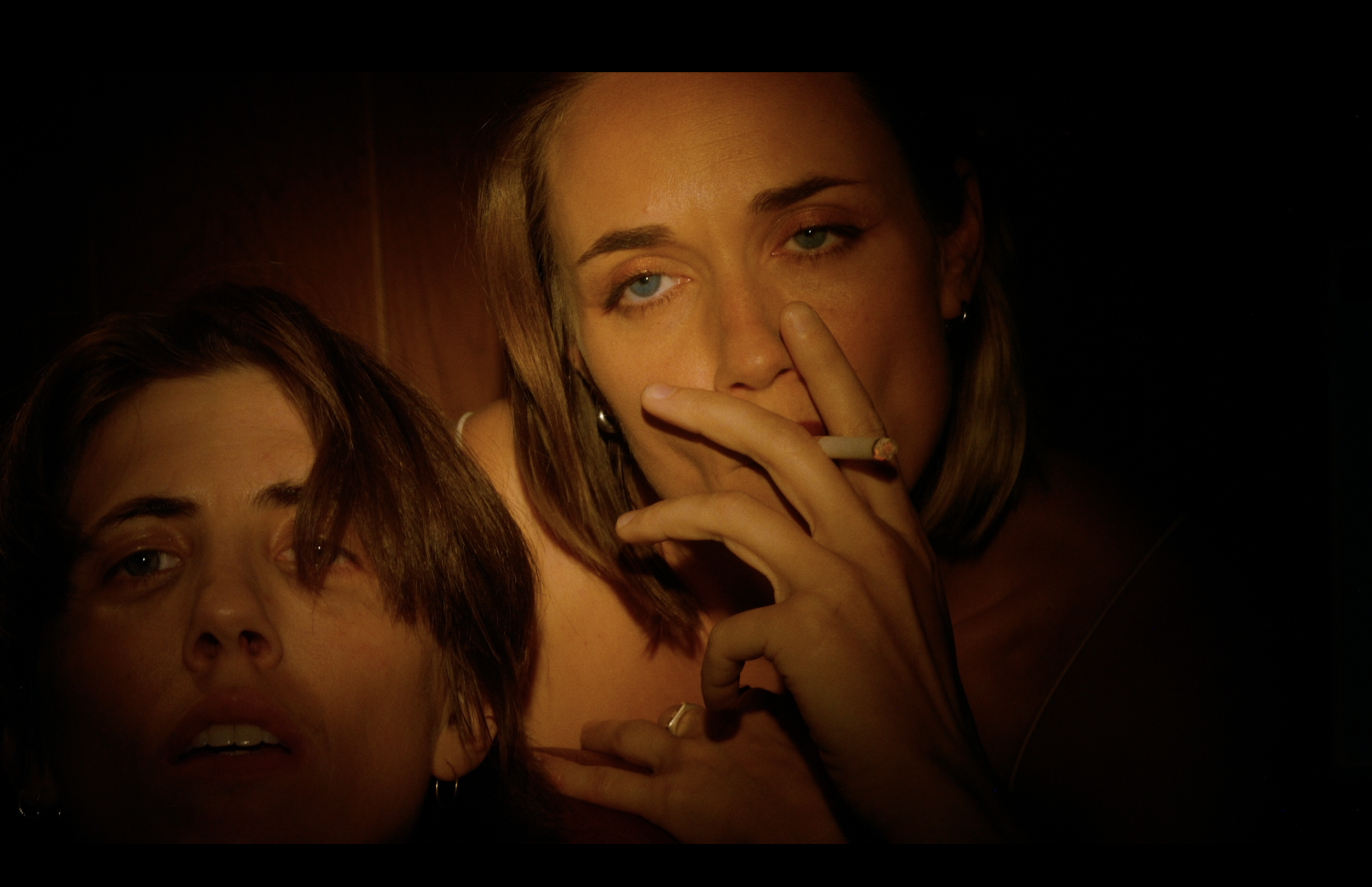 Close-up of a woman with blue eyes holding a cigarette near her nose, with another woman partially visible on the left.