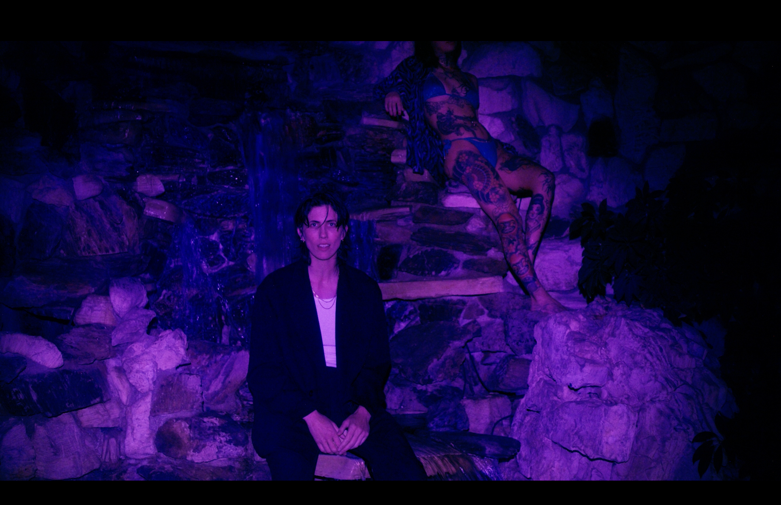 Two women in a dimly lit, purple-hued setting with stone walls. One woman in a black jacket with short dark hair is seated in front. The other woman, with tattoos and long dark hair, is sitting on a rock, wearing a blue lingerie set and a zebra print