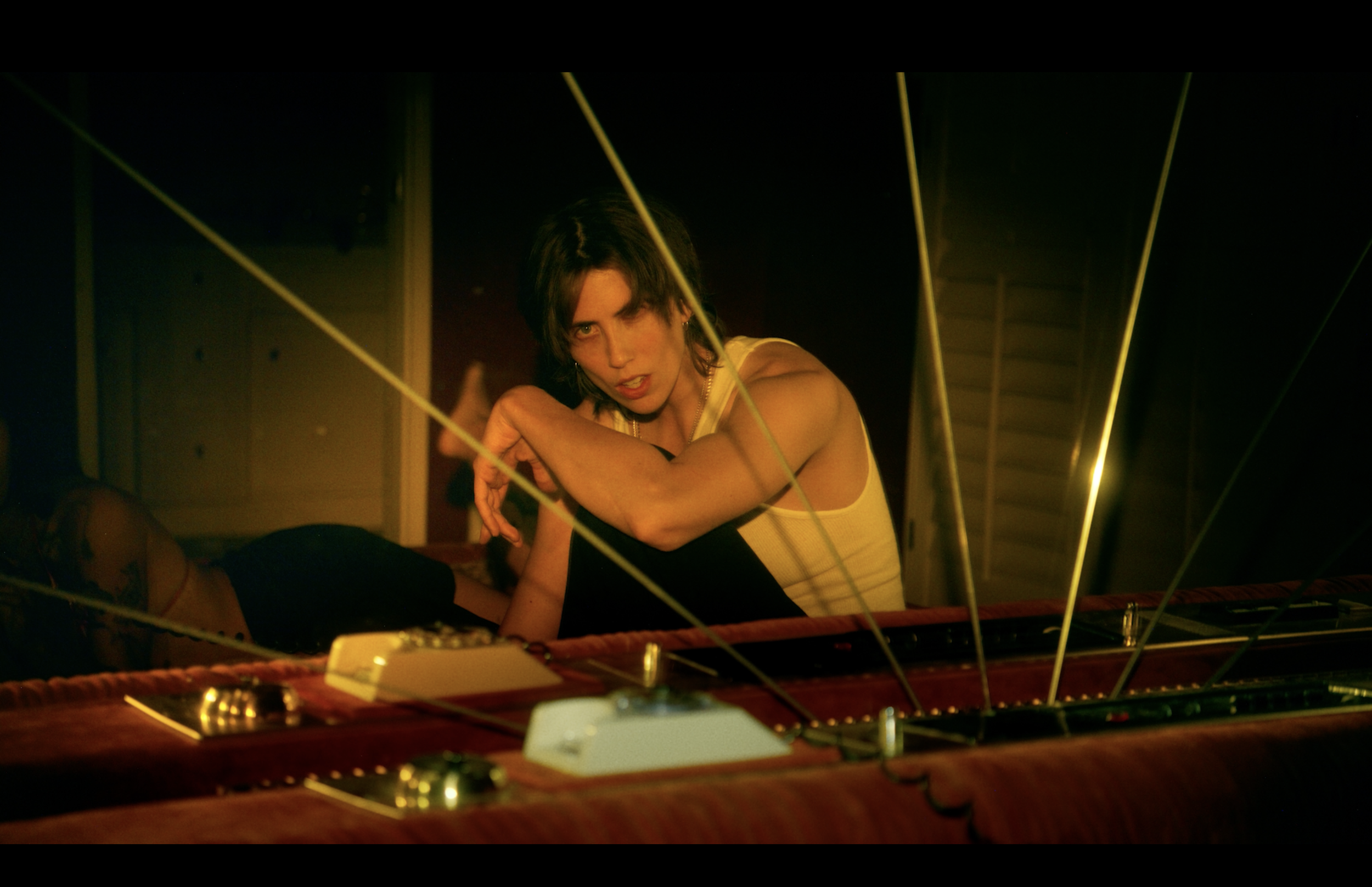 A woman with short dark hair wearing a yellow top, sitting on a bed or couch, leaning forward with her arms crossed, surrounded by yellow and green laser beams or lights, in a dimly lit room.
