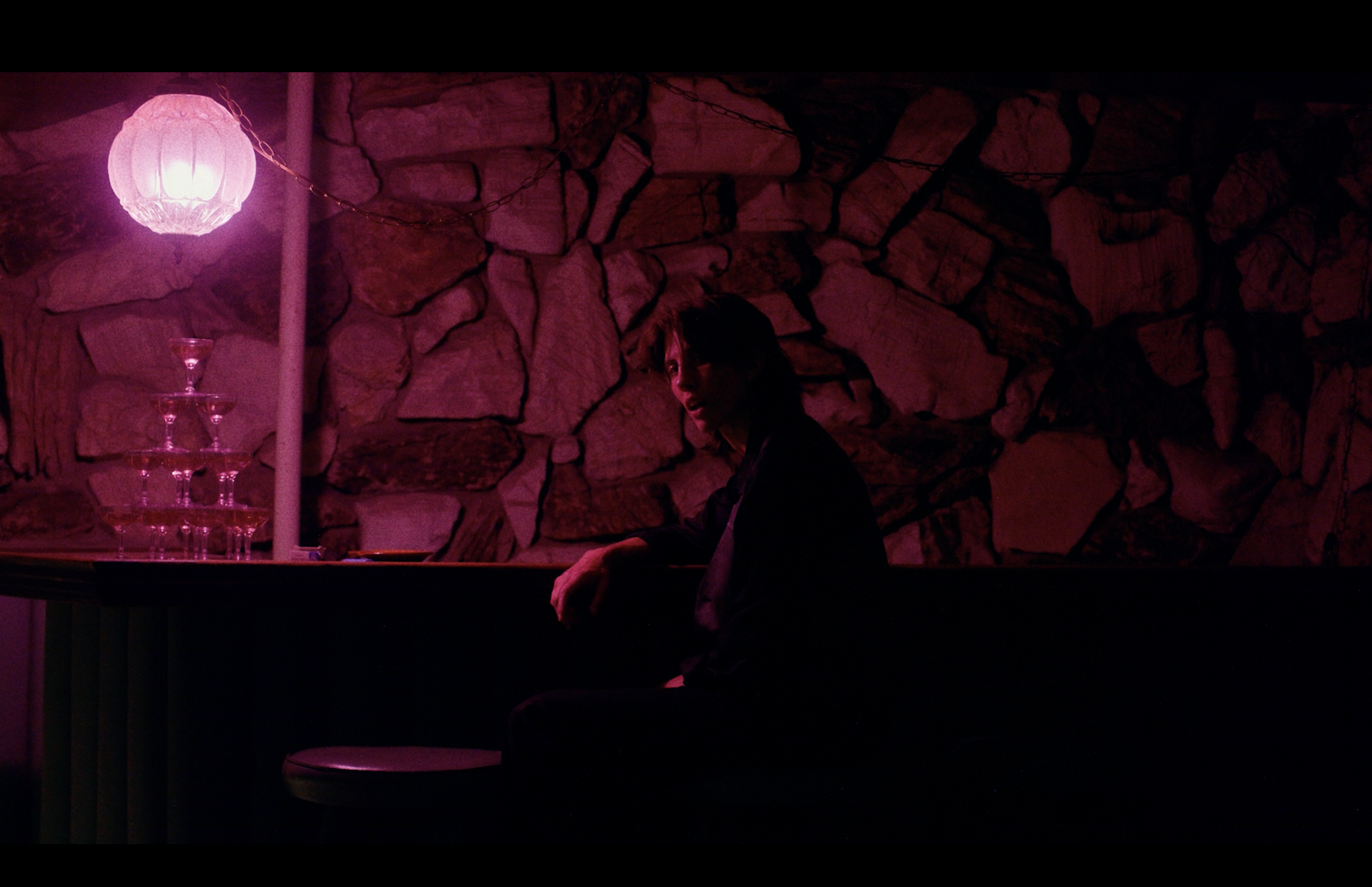 A person with dark hair sitting near a stone wall, illuminated by pink and purple lighting, with a stack of champagne glasses on a table and a pink paper lantern hanging from the ceiling.