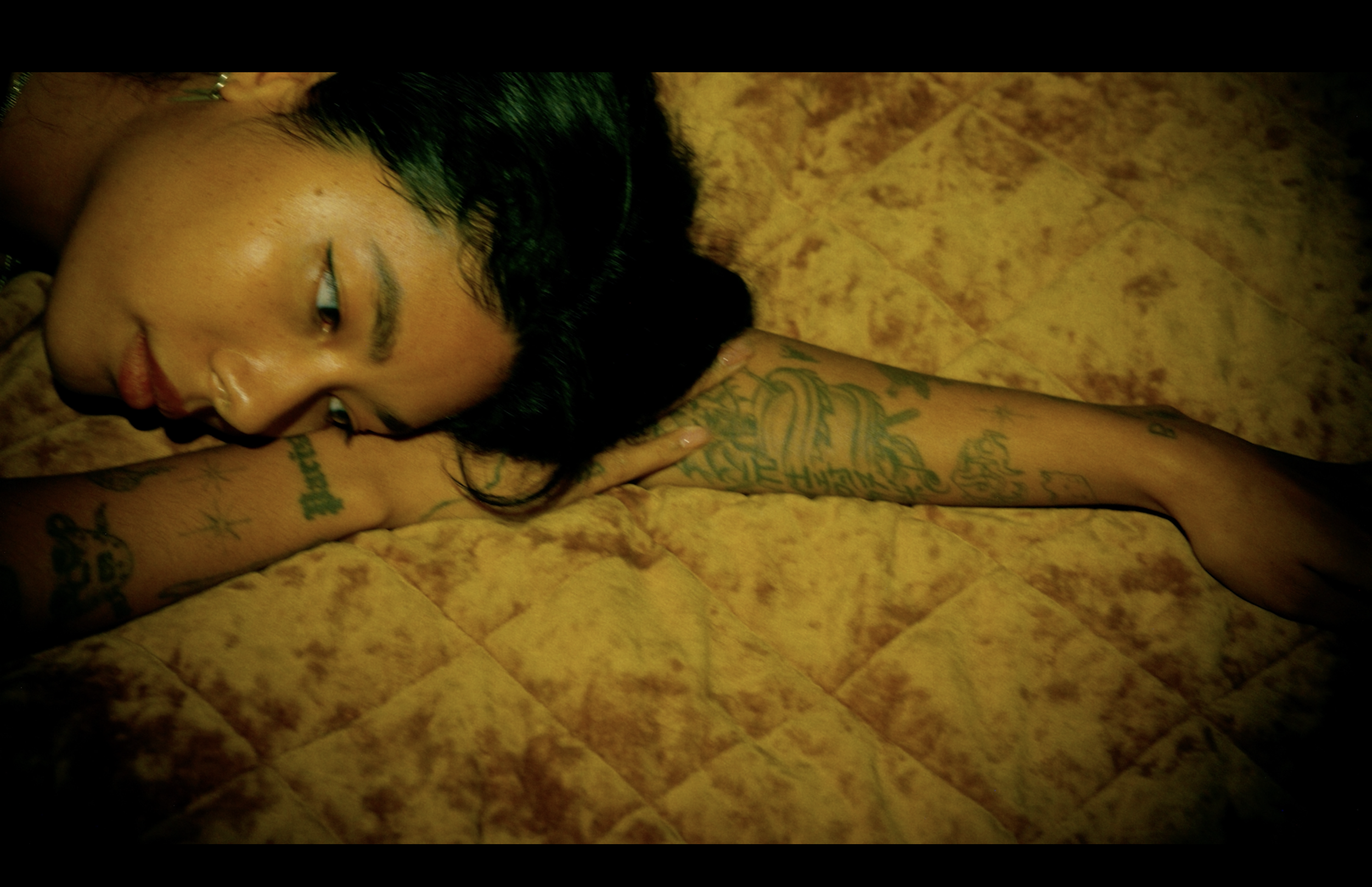 A woman with dark hair lying on a yellow textured surface, with tattoos on her arm, looking to the side.