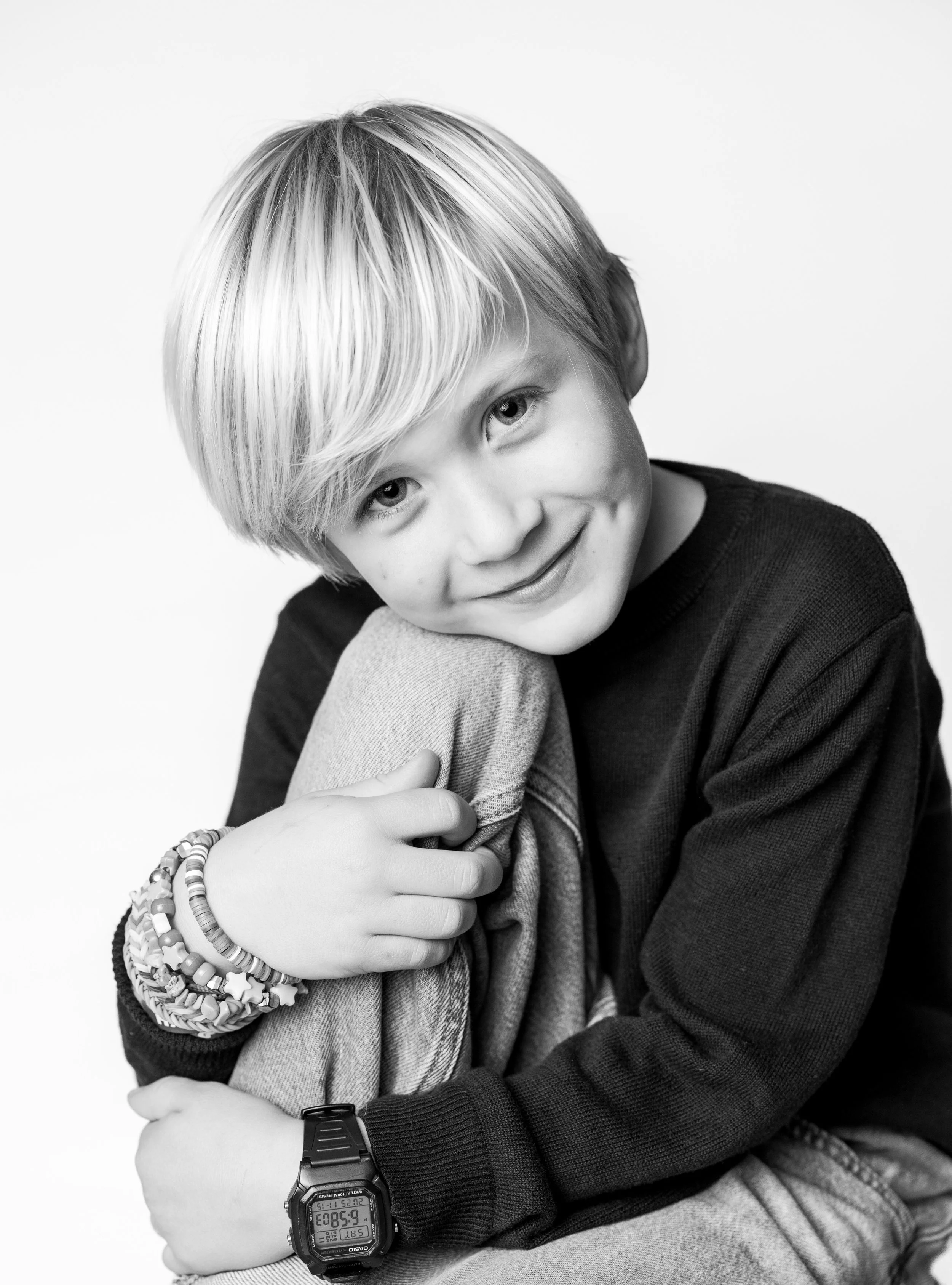 A boy with blonde hair, wearing a black long-sleeved shirt and a beaded bracelet, smiling while hugging his knee. He is wearing a digital watch on his wrist, and the background is plain.