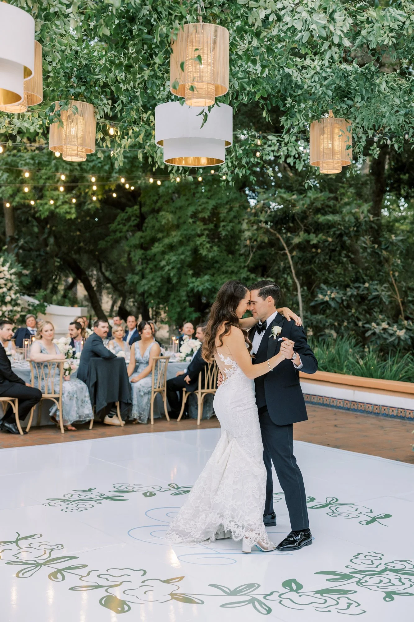 A bride and groom are dancing at their wedding reception outdoors under green trees and hanging lanterns. The bride is wearing a white lace wedding gown and the groom is in a black tuxedo. Guests are seated at tables behind them, observing the dance.