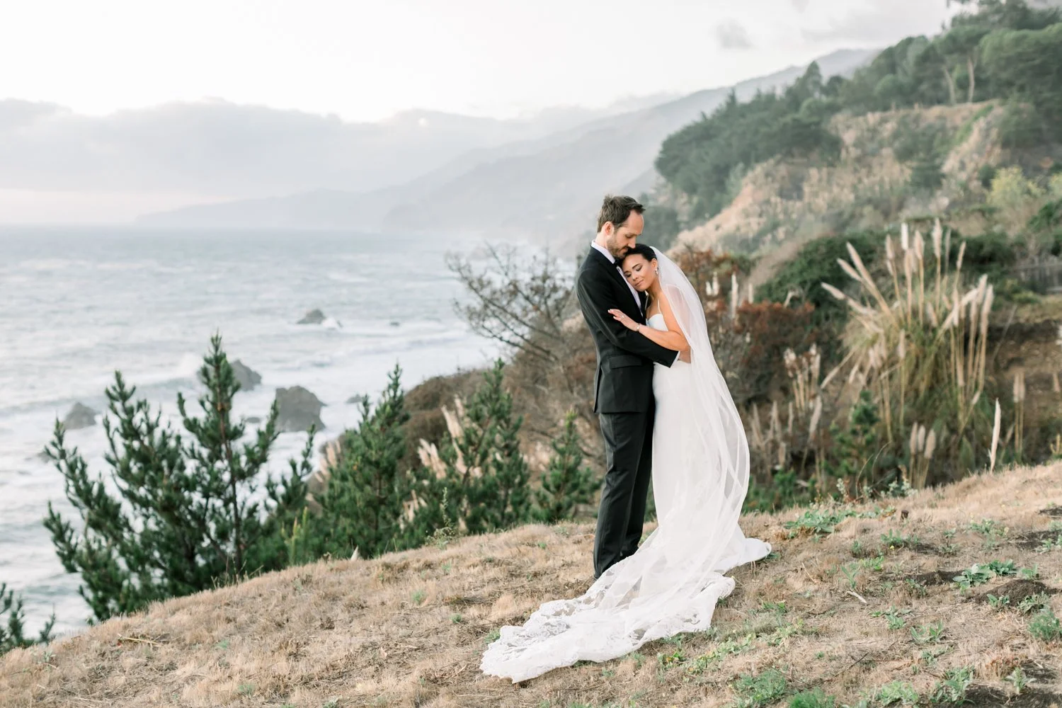 A bride and groom standing on a grassy hill near the ocean, embracing each other with the bride resting her head on the groom's chest, overlooking a rocky coastline and cloudy sky.
