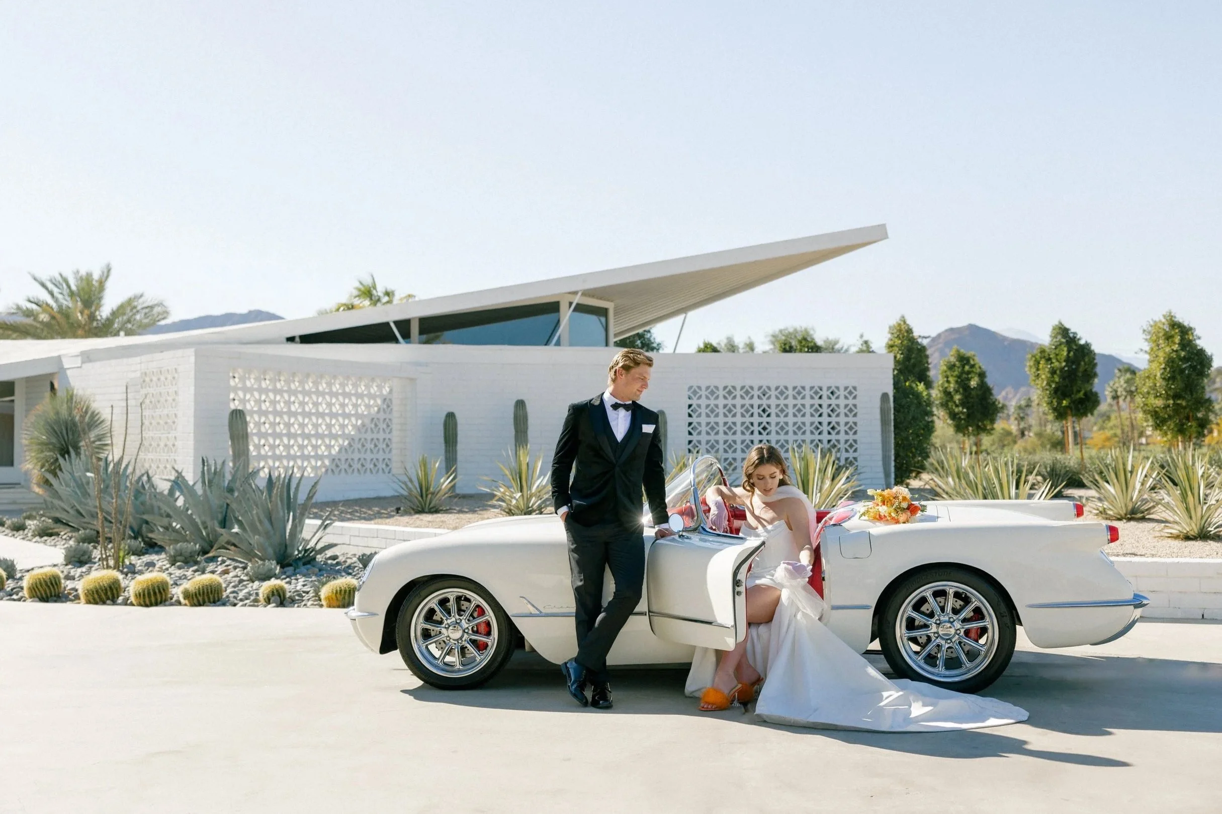 A woman in a wedding dress sitting in a vintage white convertible car, with a man in a tuxedo standing beside her, in a modern outdoor setting with desert plants and a white building in the background.