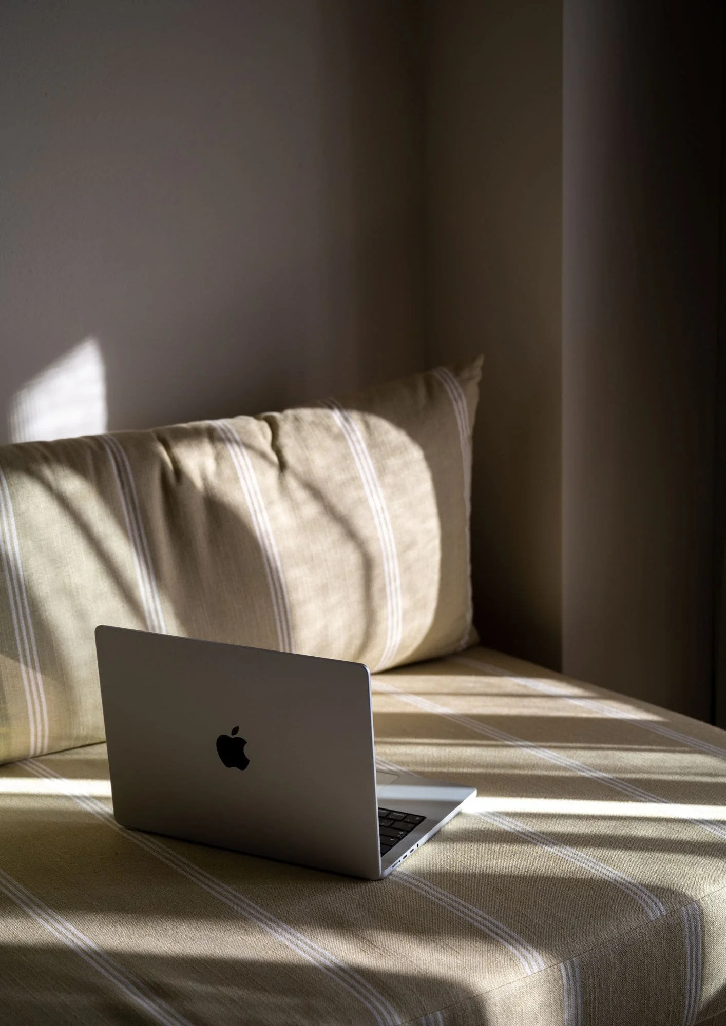 A MacBook laptop on a beige, striped tablecloth, with a beige striped pillow on a bed or couch behind it, in a room with beige walls and sunlight casting shadows.