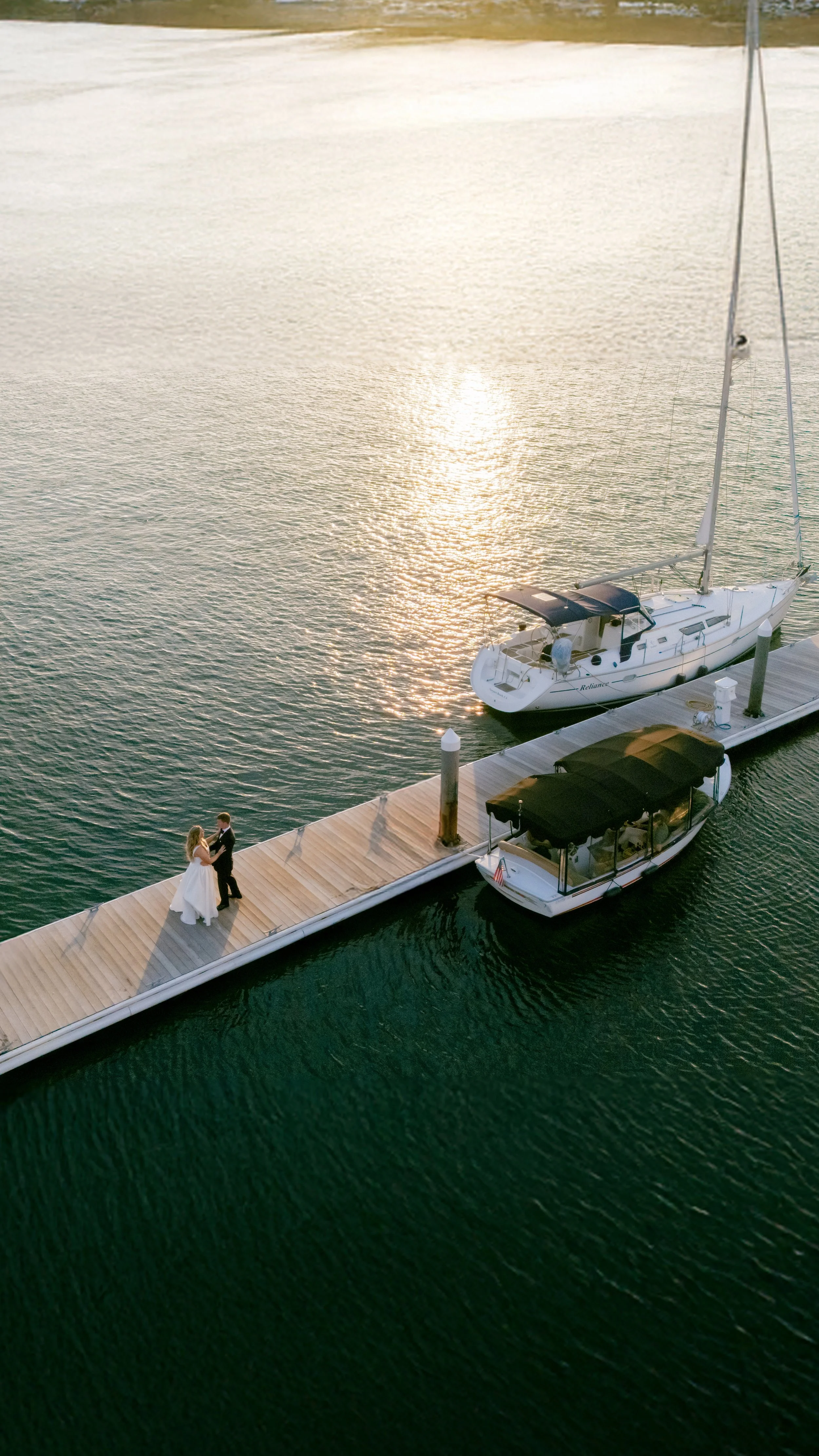 A bride and groom standing on a wooden dock beside two boats on a body of water. The water reflects the light of the setting or rising sun.