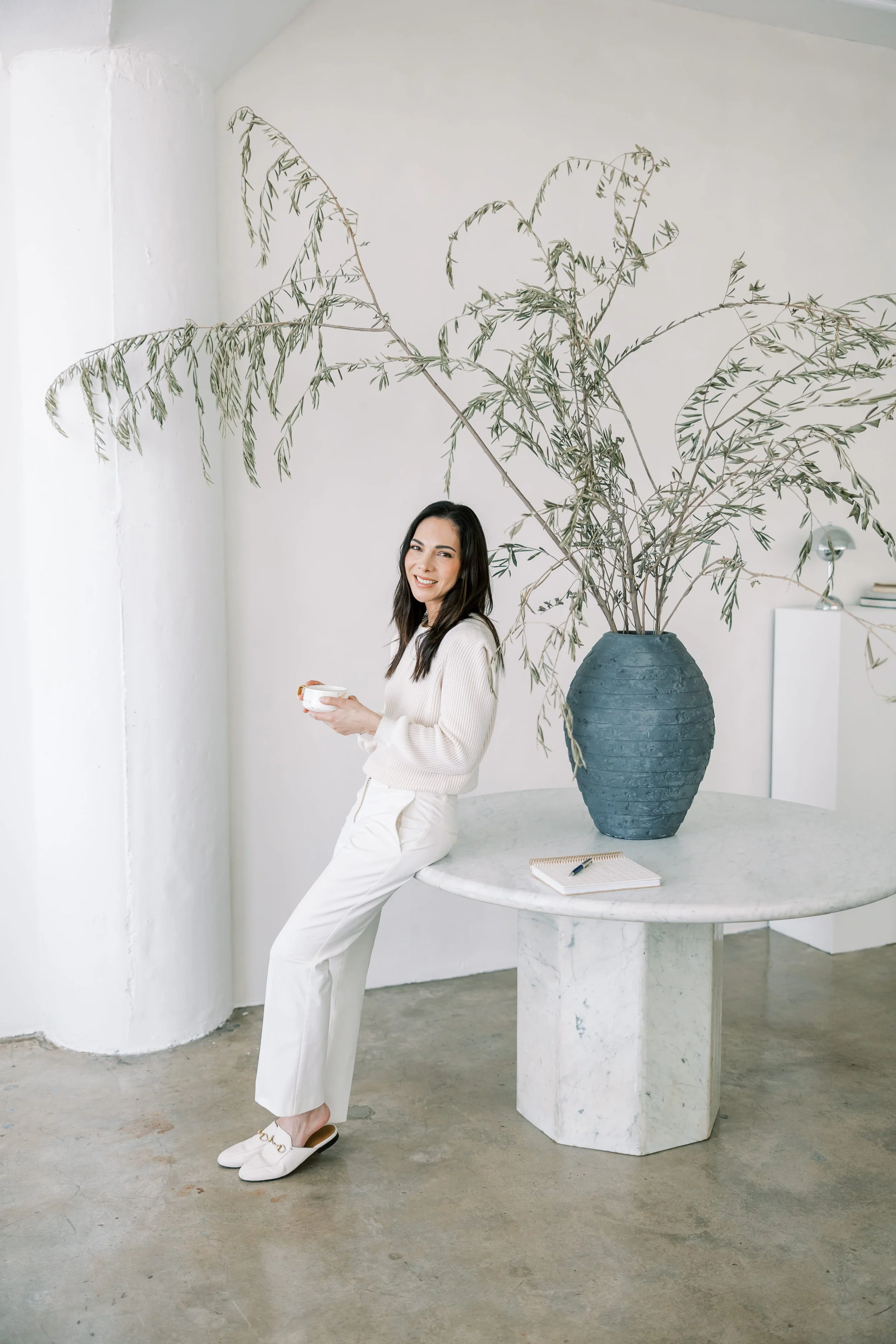 A woman with long dark hair, smiling, stands leaning against a round marble table in a bright, minimalist room. She is holding a small white cup, dressed in a cream sweater and white pants with white shoes. A large, blue ceramic vase with tall, leafy branches is on the table, with a notebook and pen placed beside it. The room has white walls and a concrete floor.