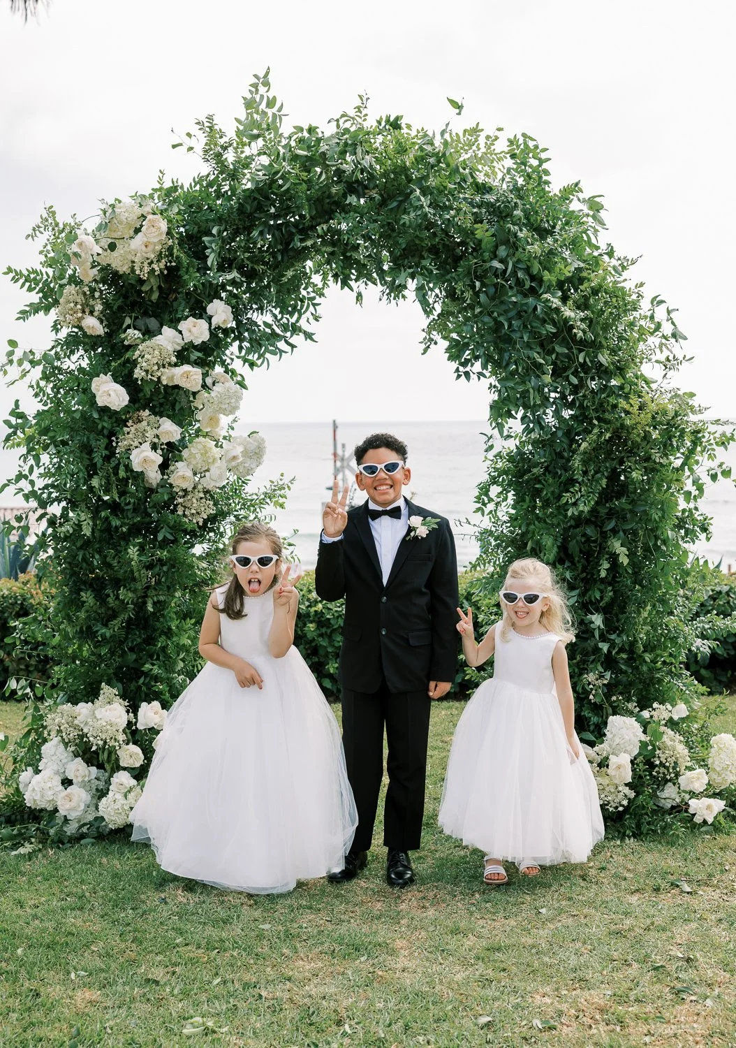 Ring bearer and two flower girls make a silly face for the camera while standing in front of the ceremony arch at Ole Hanson beach club in san clemente. 