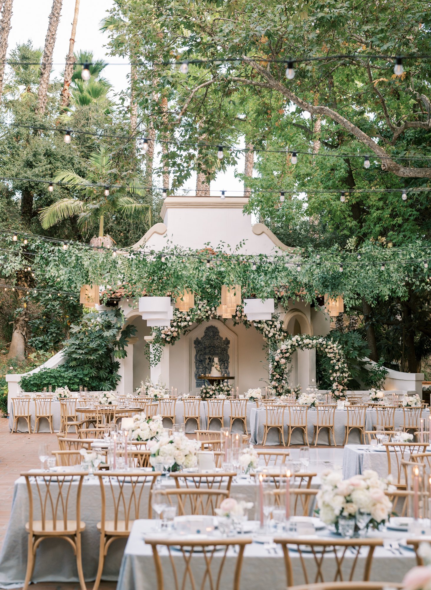 Elegant outdoor wedding reception setup with tables, chairs, and floral arrangements under string lights and trees.