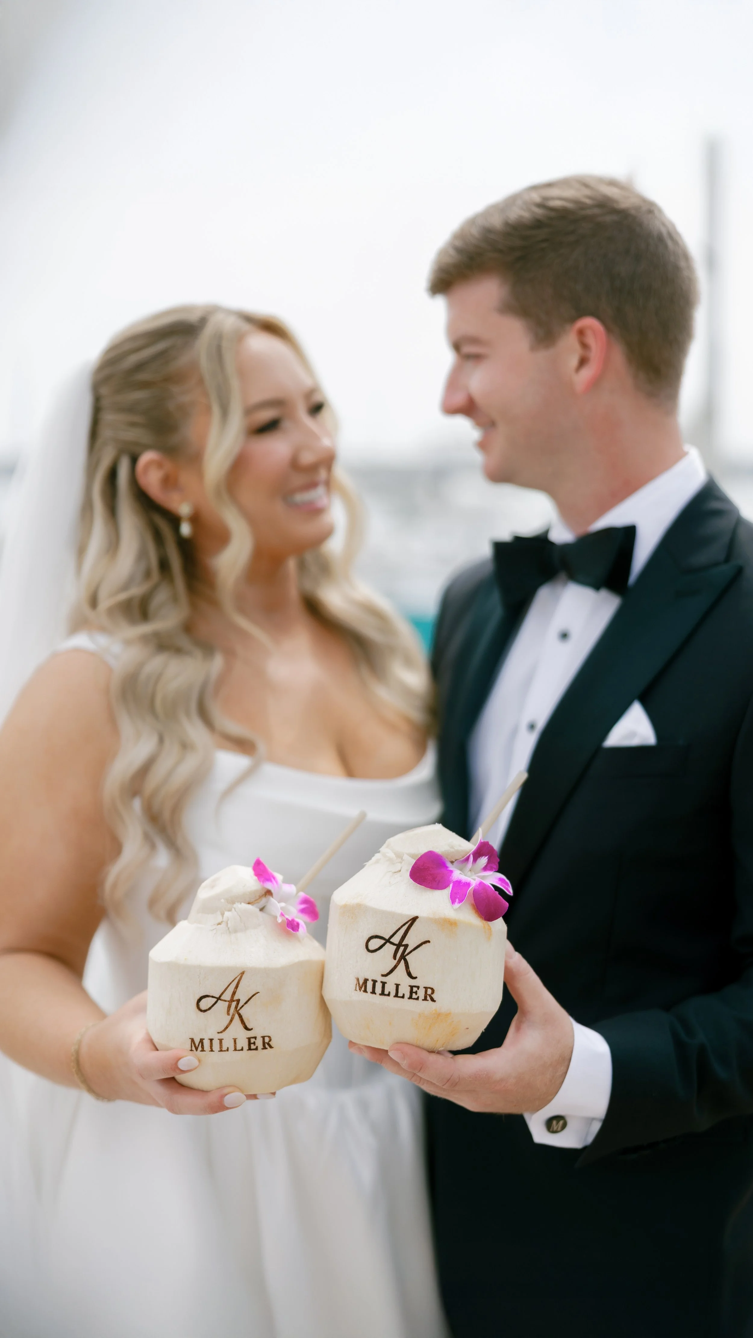 A bride and groom holding coconuts decorated with pink orchids and personalized with the names 'Miller' and the initials 'AK'. The bride is wearing a white wedding dress with long blonde hair, and the groom is wearing a black tuxedo with a bow tie.