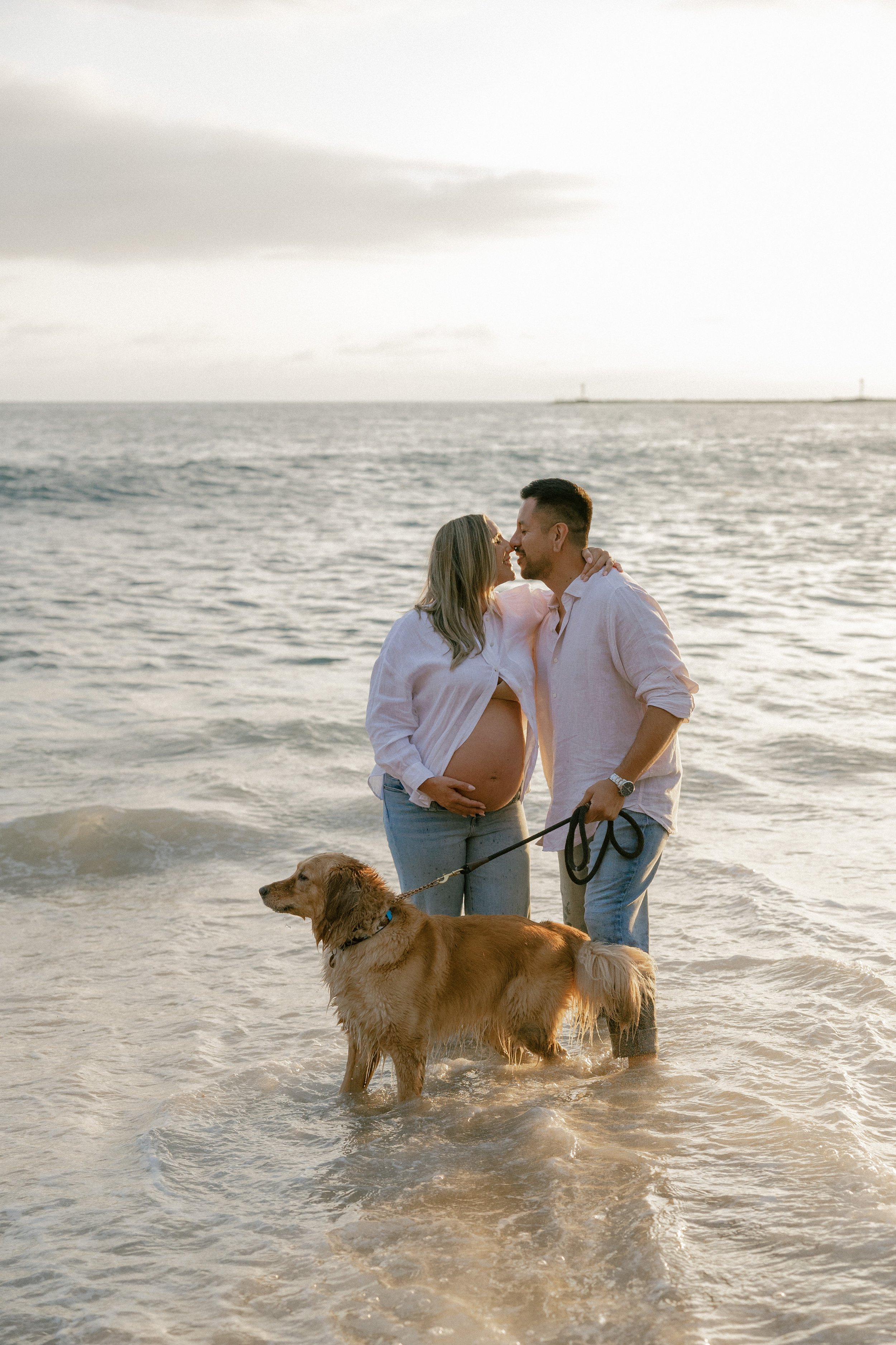 A pregnant woman and a man are standing on the beach in shallow water, with the woman gently touching her belly. They are smiling and about to kiss. There is a golden retriever dog in the foreground, on a leash, standing in the water. The background shows the ocean with a cloudy sky and some distant structures on the horizon.