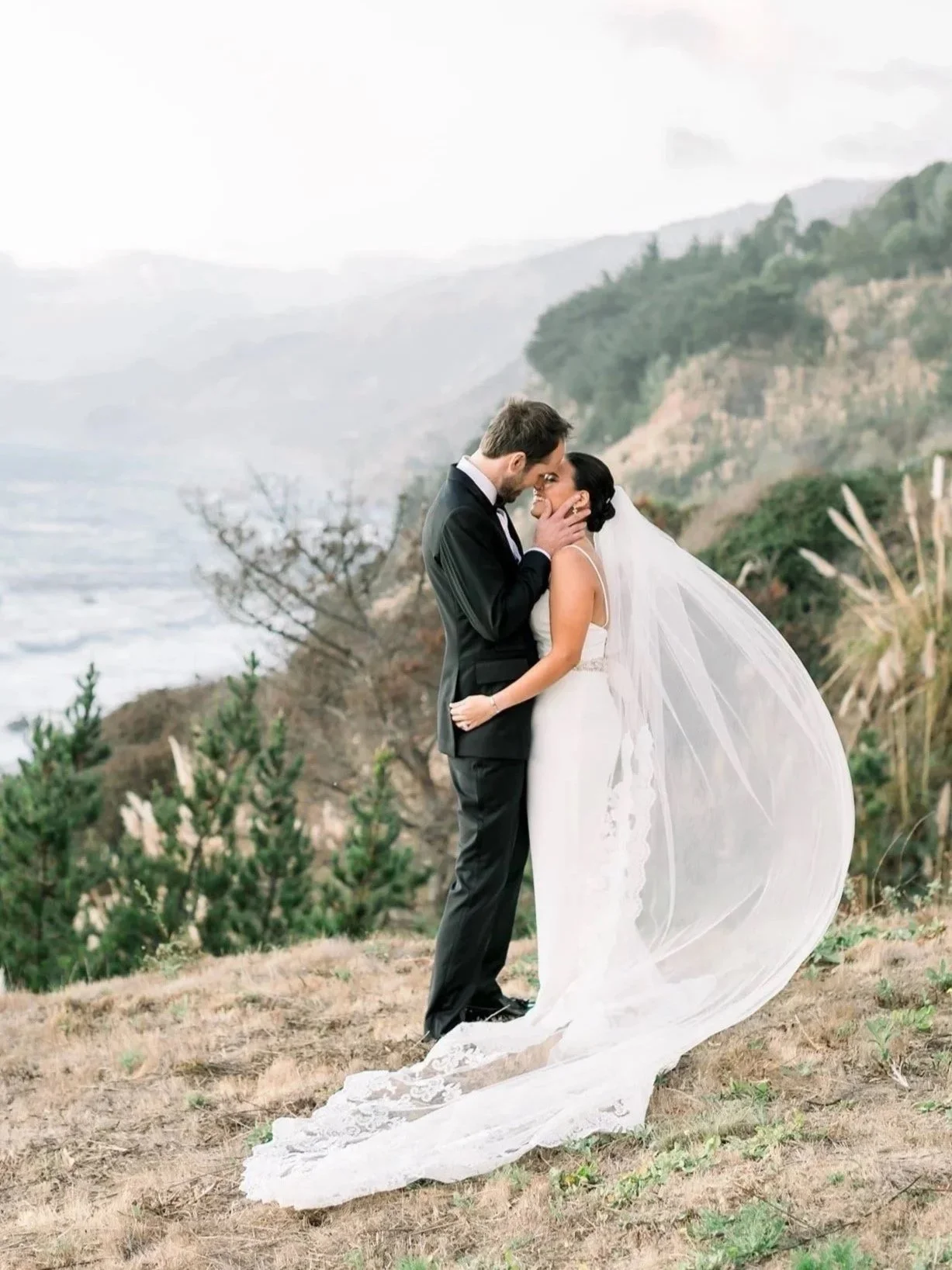 A bride and groom sharing a kiss outdoors on a hillside with a scenic mountain and ocean view in the background, the bride wearing a white wedding gown with a long veil and the groom in a black suit.