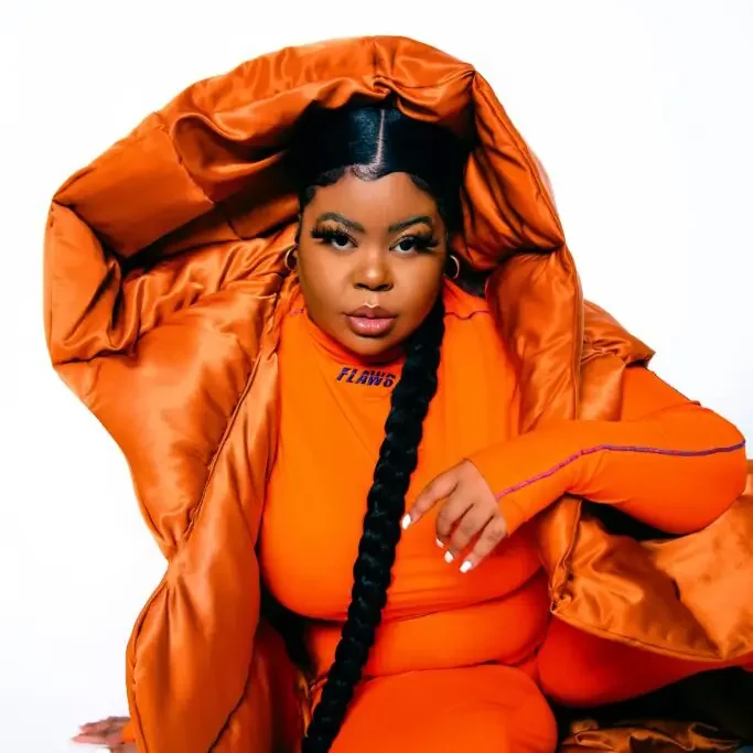 A woman with long black braided hair, wearing an orange puffer jacket and matching orange outfit, sitting against a plain white background.