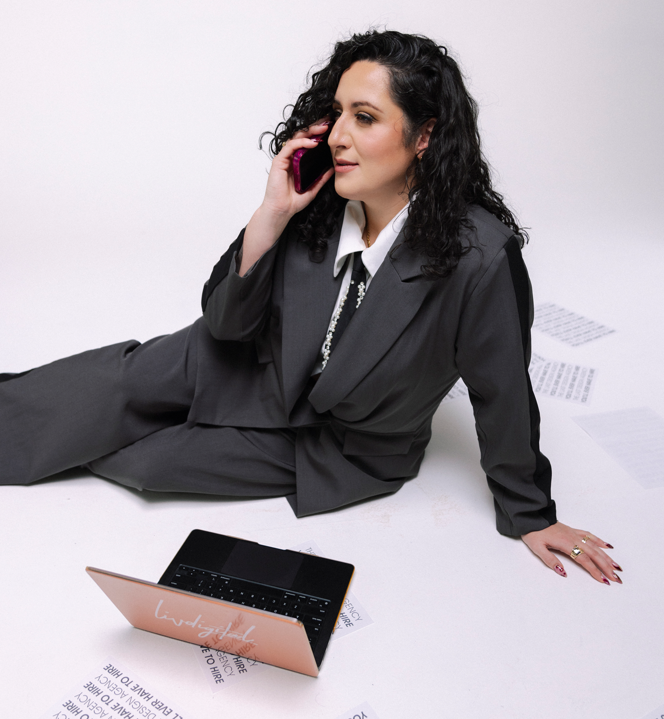 A woman in a black suit sitting on the floor, talking on a cell phone, with papers and a laptop around her.