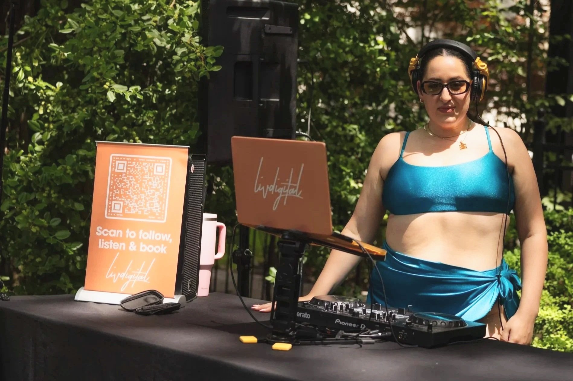 A woman DJ wearing sunglasses, a blue crop top, and a matching wrap skirt, standing outdoors at a DJ setup with a laptop, mixer, and sign, surrounded by greenery.