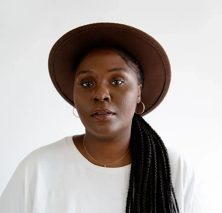 A woman wearing a brown wide-brimmed hat, white top, gold hoop earrings, and a delicate gold necklace, looking directly at the camera against a plain white background.