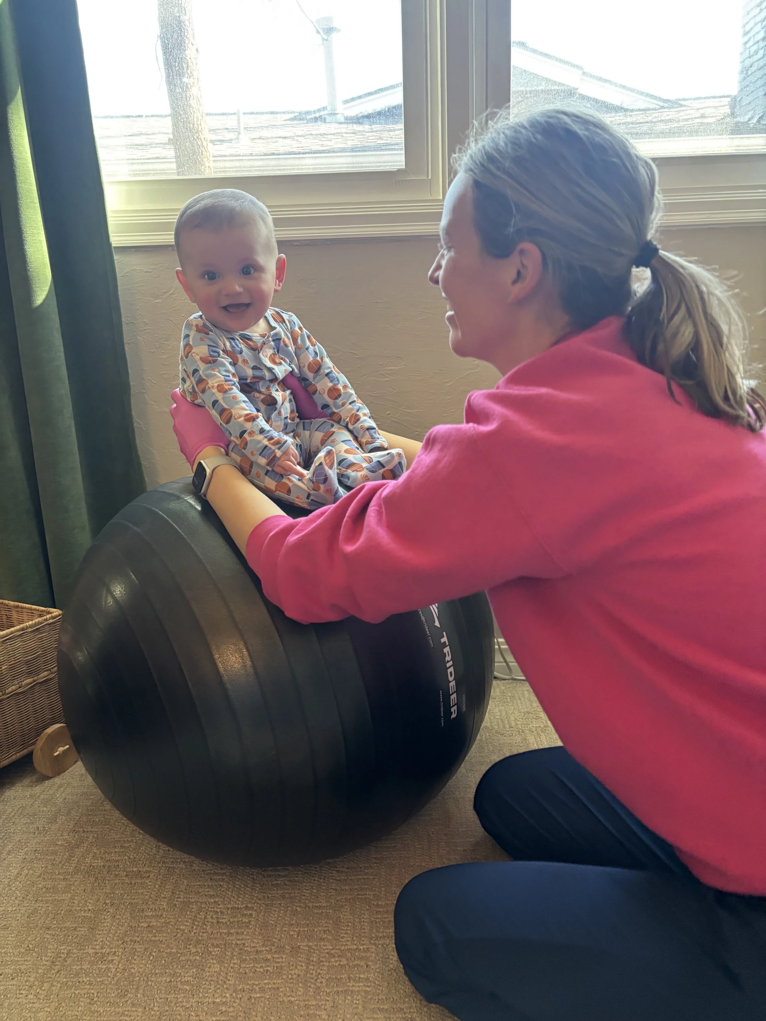 A physical therapist supporting a baby in a patterned onesie and smiling, with a large exercise ball to work on sitting balance while making it a fun experience for baby.