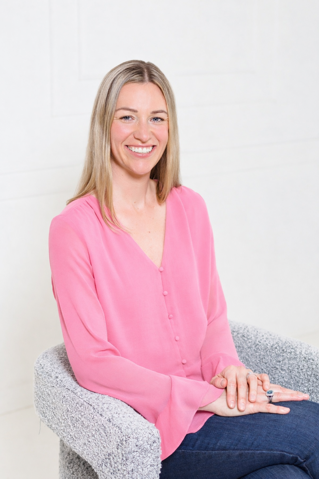 A woman with blonde hair wearing a pink blouse and dark jeans sitting on a textured chair against a white background, smiling.