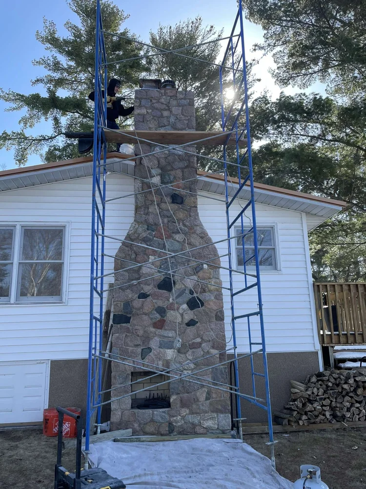 Scaffold up on a tall stone chimney, tarp down to keep the yard clean.