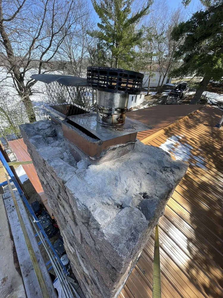 An old crown with a stainless flue cap on a stone chimney, metal roof below