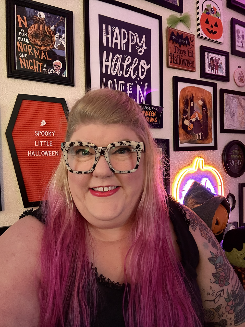 Smile of a woman with pink hair and patterned glasses taking a selfie in a Halloween-themed room decorated with various Halloween posters and signs