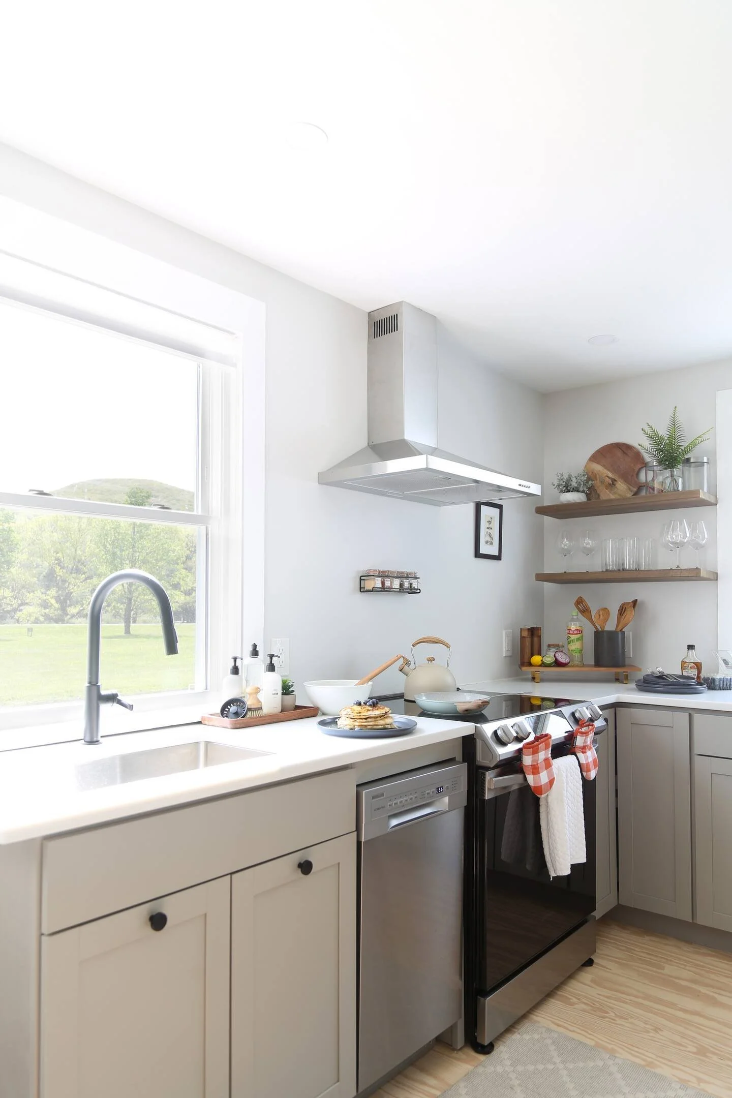 Modern kitchen with white cabinets, black knobs, stainless steel appliances, open shelves with glassware, cutting boards, and potted plants, and a window showing green trees outside.