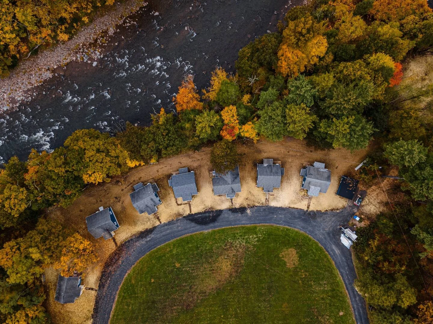 An aerial view of eight houses in a semi-circle at the edge of a green golf course, surrounded by trees with autumn foliage, and a river running nearby.