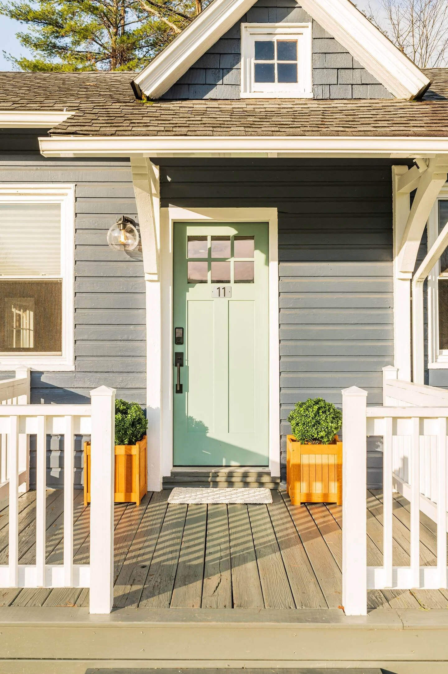 Front porch of a house with a light green door, two potted bushes, and a white fence, with a gray house exterior and small window above the door.