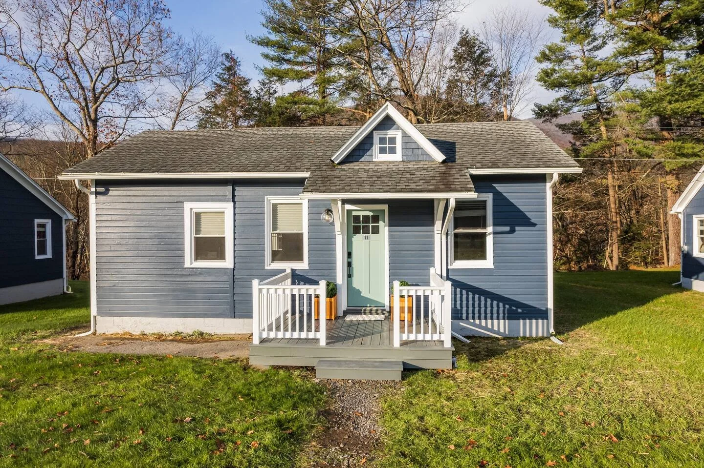 Blue house with white trim, small front porch, green door, and two small planters on each side, surrounded by green grass and trees.