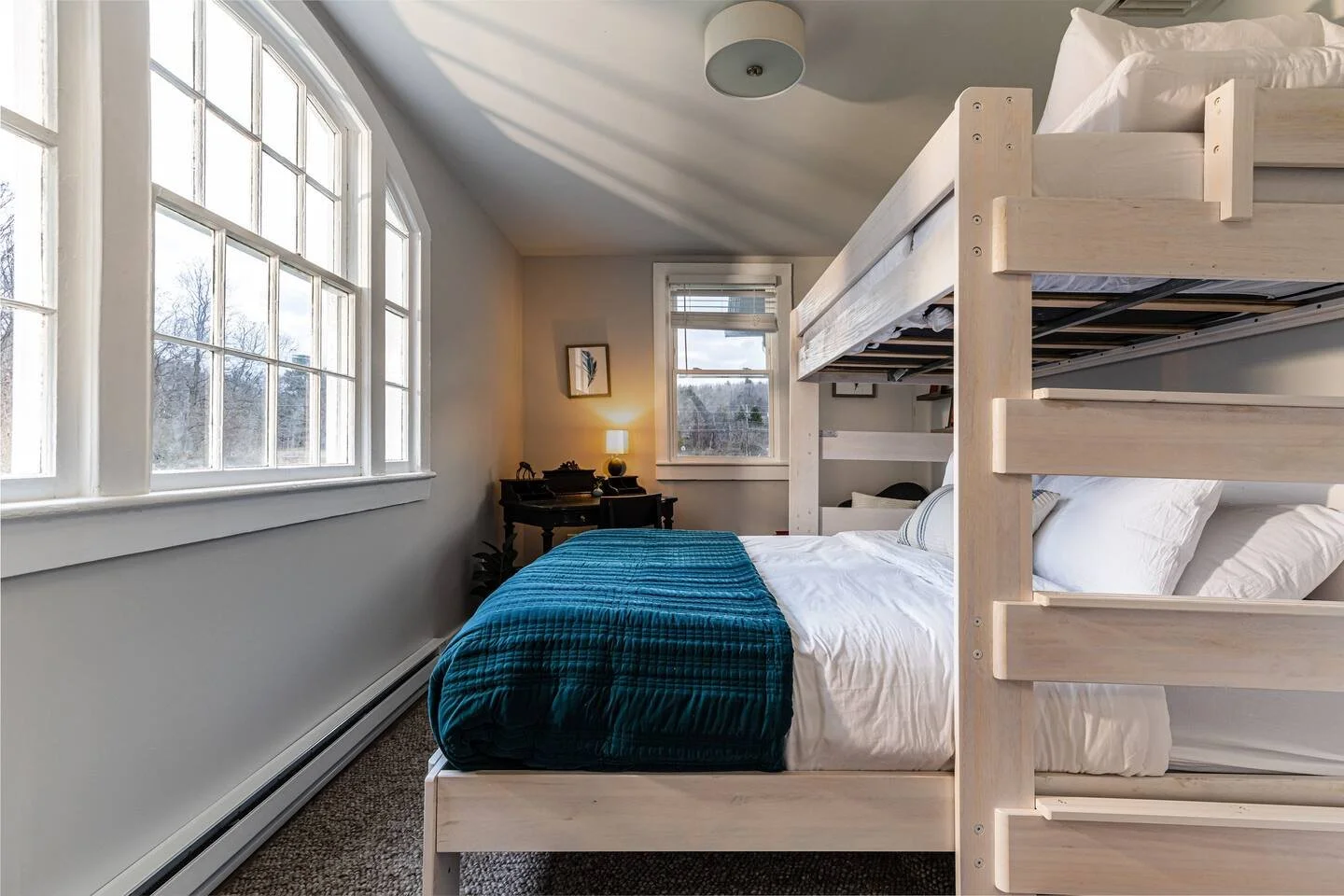A cozy bedroom featuring a bunk bed with white bedding, a blue blanket, a nightstand with a lamp, a window letting in natural light, and a small wall art piece.