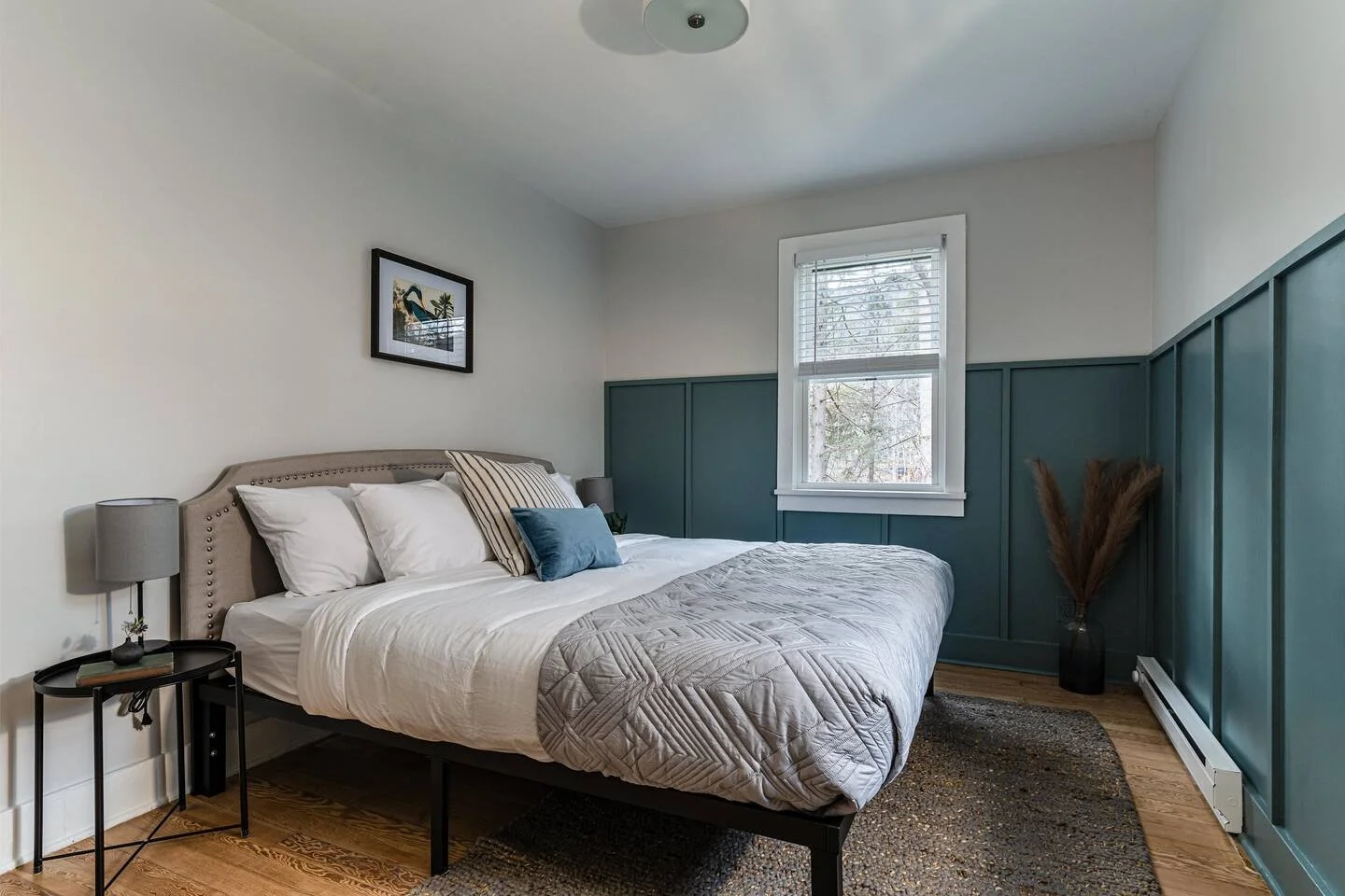 Bedroom with a beige upholstered headboard, white bedding, and decorative pillows, positioned beside a black side table with a gray lamp. The room has a window with blinds, wood flooring, a textured rug, and an accent wall painted teal with a black vase containing dried grasses.