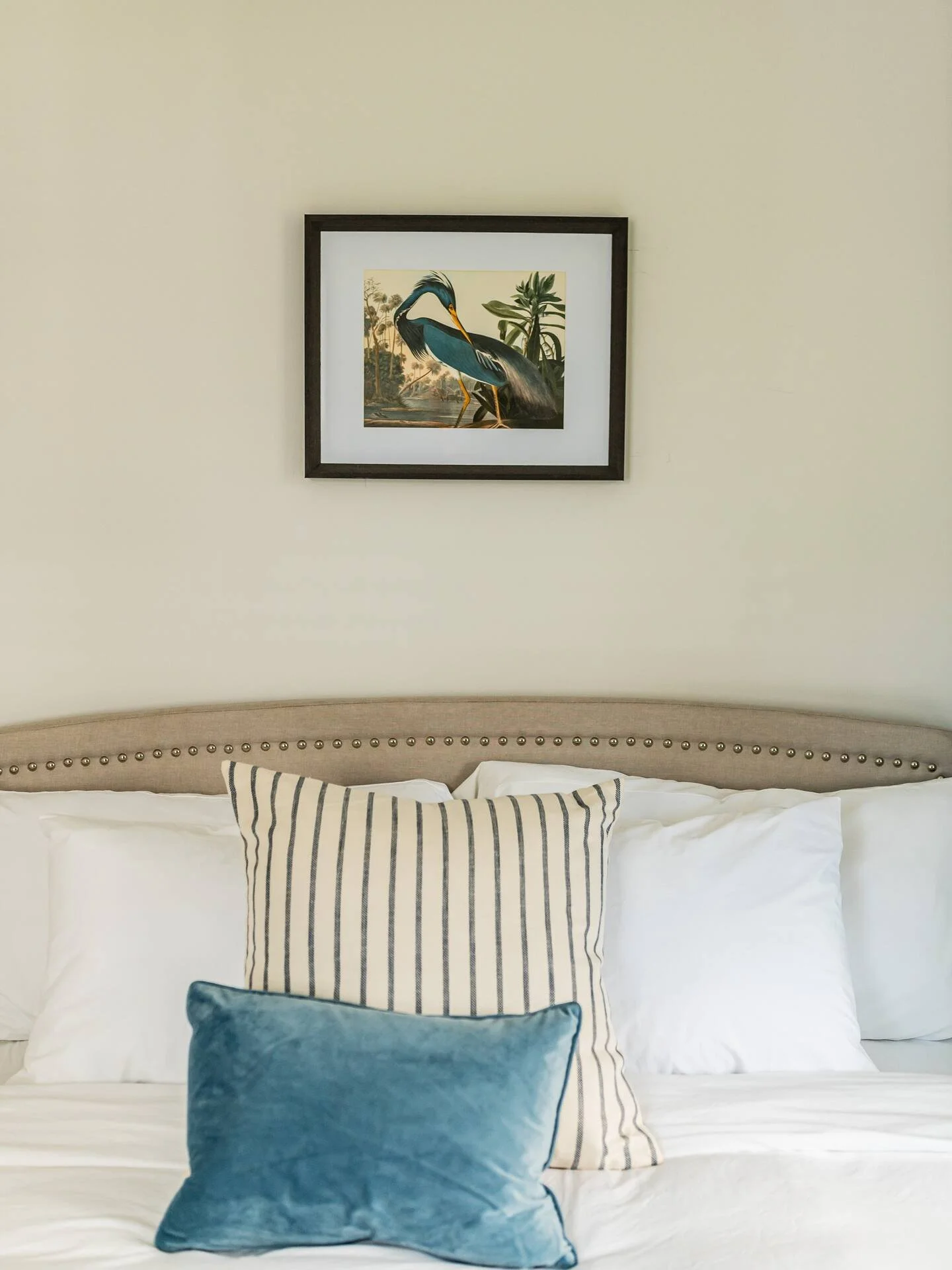 A bed with multiple pillows in front of a beige wall, with a framed picture of a heron and plants hanging above.