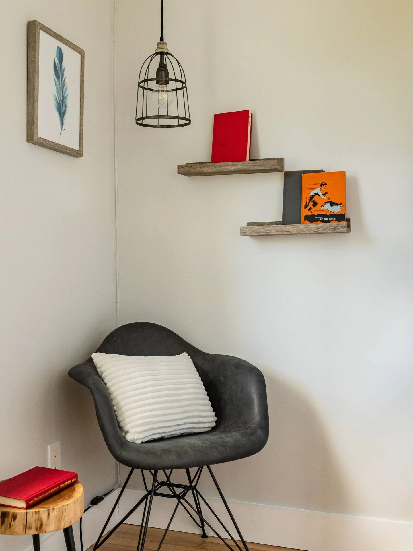 A cozy corner with a black modern chair, white striped pillow, small wooden side table with a red book, floating wooden shelves with red and orange books, a framed feather artwork, and a hanging black wire light fixture.
