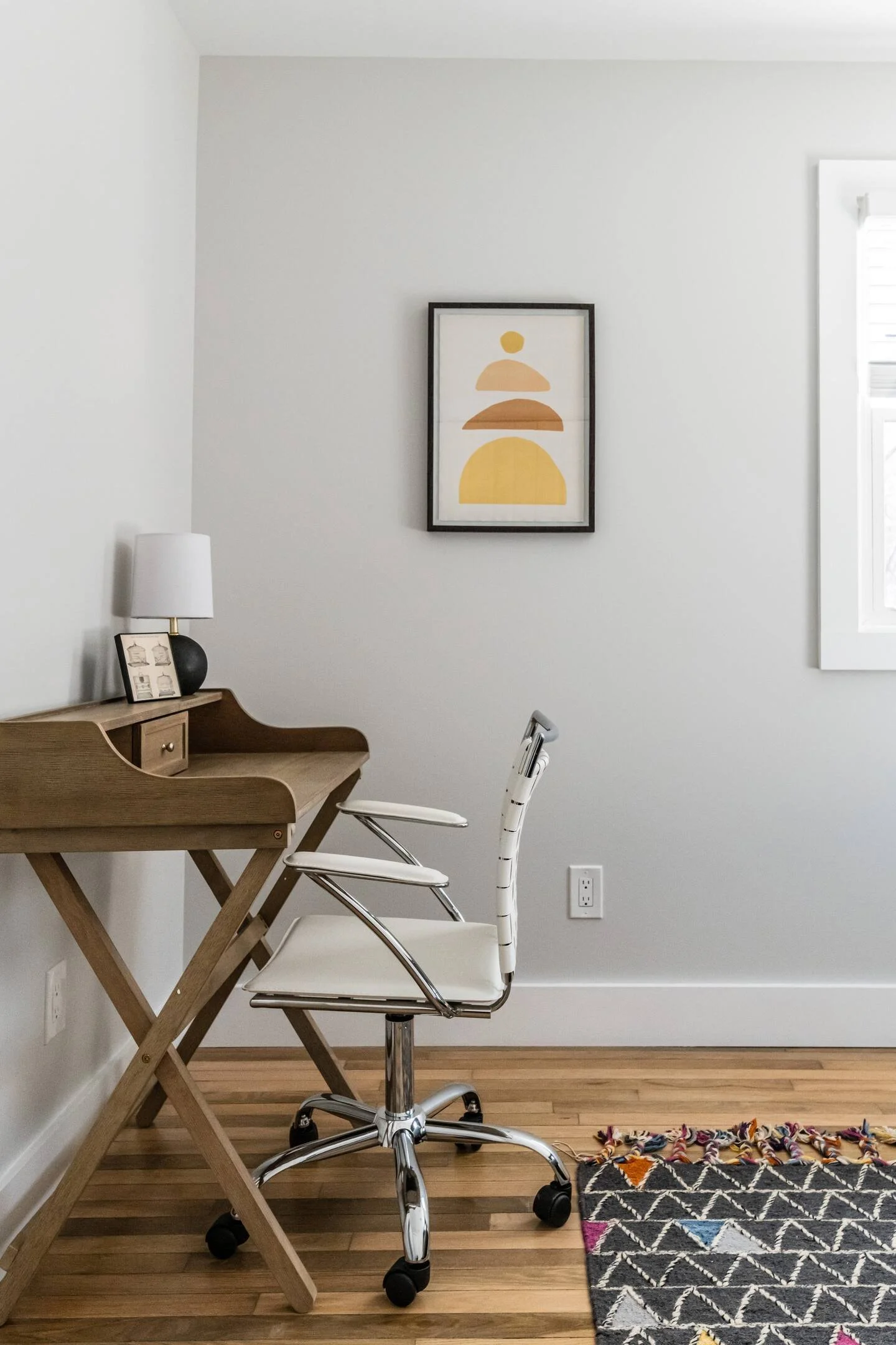A minimalist workspace with a wooden desk, white office chair, a framed abstract art piece on the wall, and a window with white shutters letting in light. A patterned rug is partially visible on the floor.