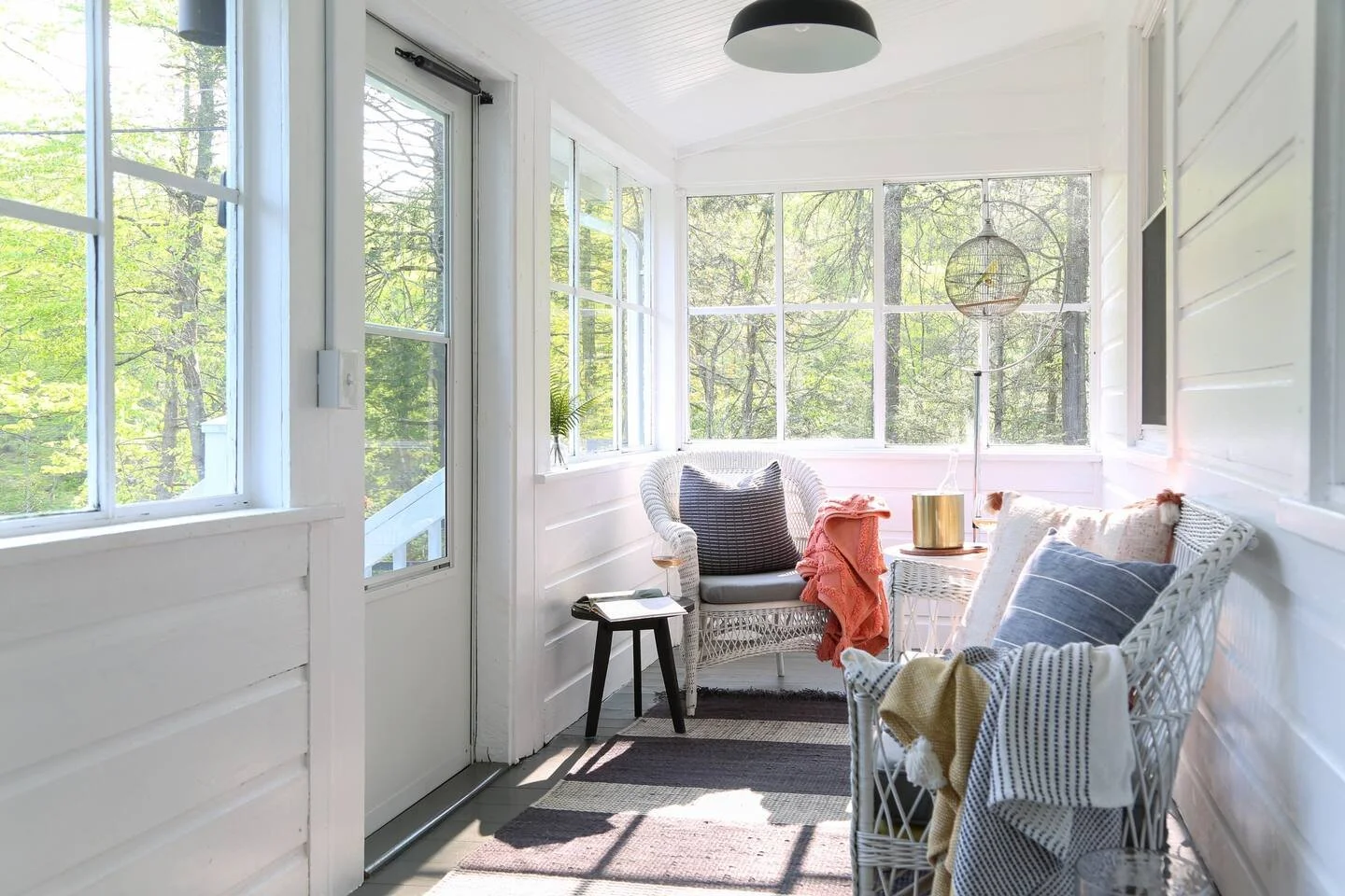 Sunlit enclosed porch with wicker chairs, pillows, a small side table, and potted plants, surrounded by green trees visible through the windows