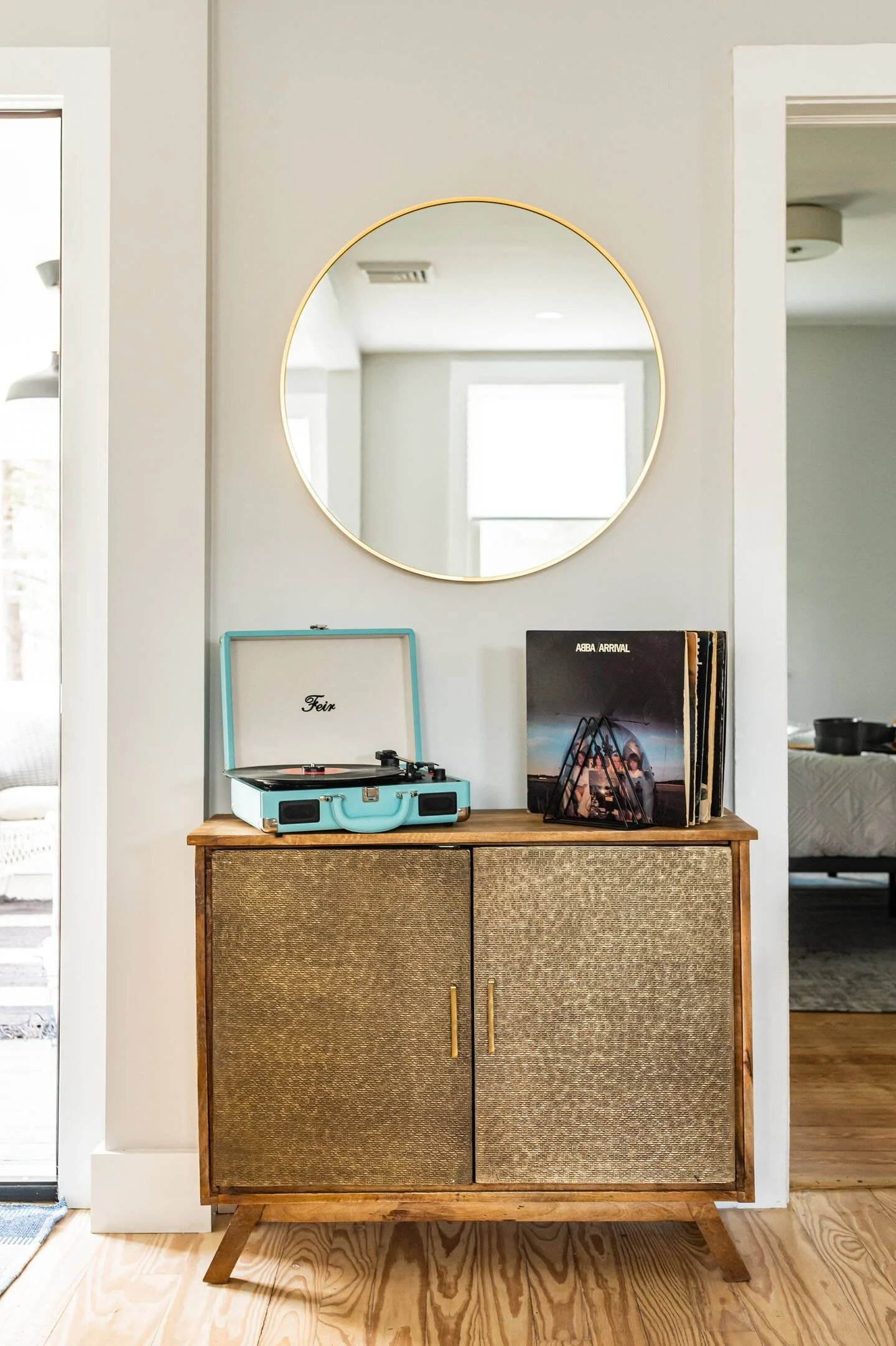A wooden cabinet with textured doors, a vintage blue record player on top, and a set of vinyl records beside it, reflected in a round mirror on a white wall.