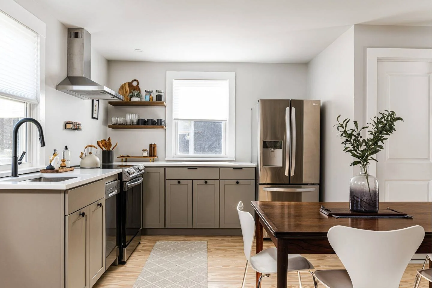 Modern kitchen with beige cabinets, stainless steel appliances, wooden dining table with white chairs, and a large vase with green foliage centerpiece.