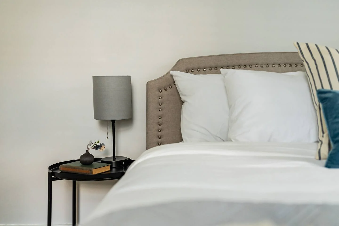 A neatly made bed with white pillows and sheets, a beige upholstered headboard with nailhead trim, a stylish black side table with a gray lampshade, a small black vase with flowers, a closed book, and a striped pillow.