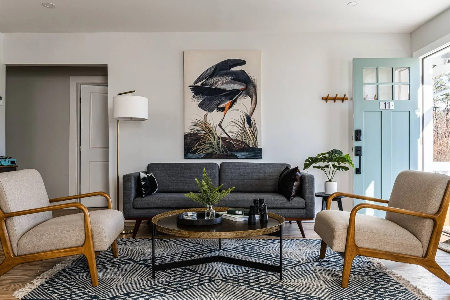 Living room with a gray sofa, two beige armchairs with wooden frames, and a round wooden coffee table. There is a large artwork of a heron on the wall, a white floor lamp, a potted plant, and a light blue front door with glass panels.