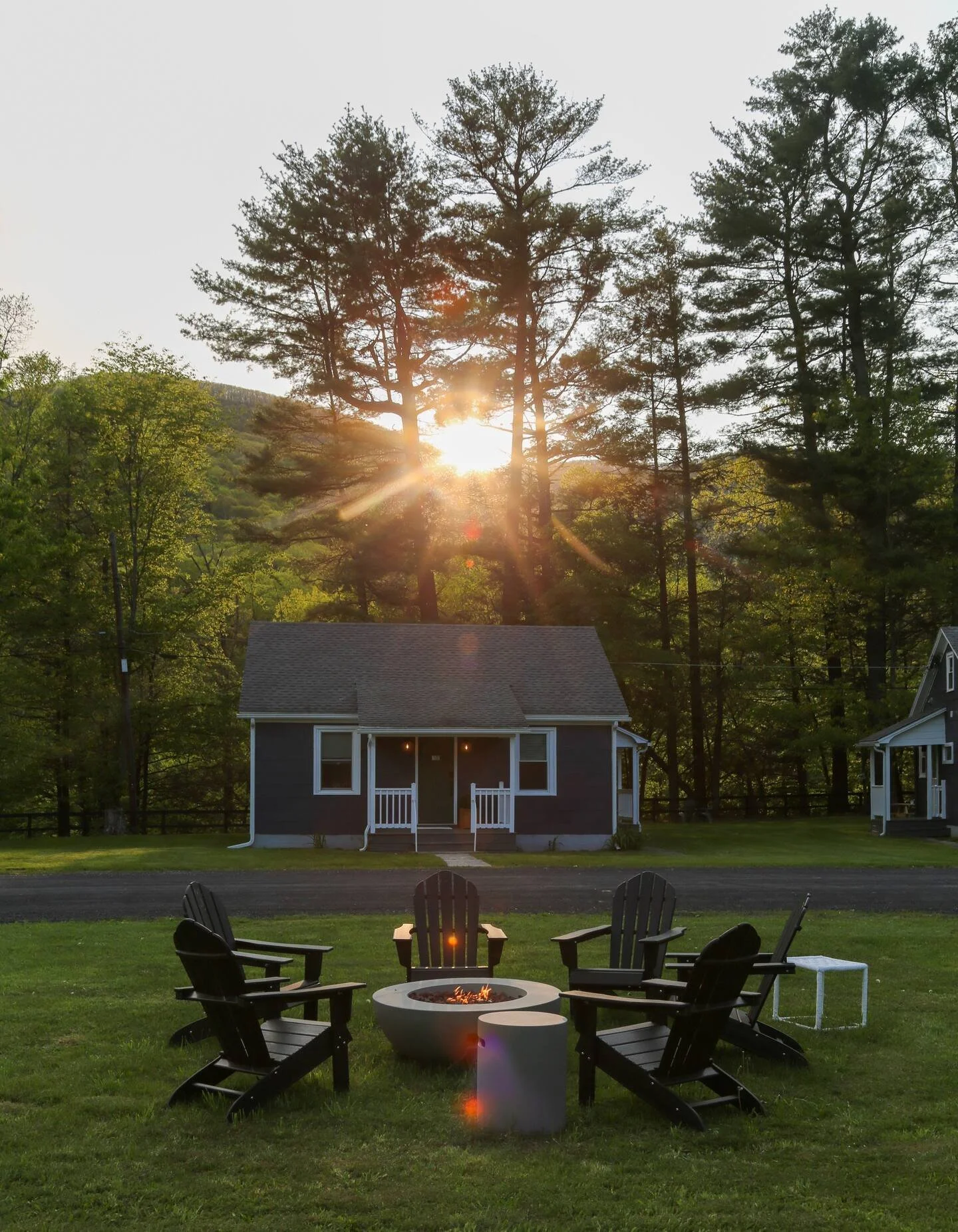 Sunset over a backyard with six black Adirondack chairs arranged around a fire pit, a small white side table, a dark house with a porch, and tall trees in the background.