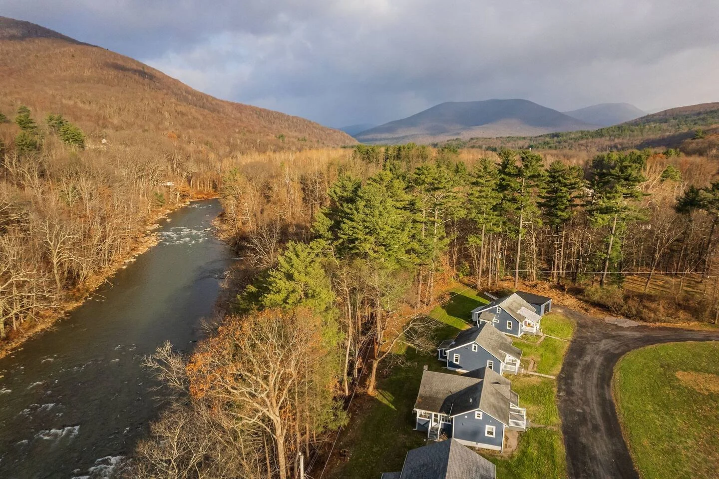 Aerial view of a rural landscape with a river running through a forested area and a small group of houses nearby, with mountains in the background during daytime in late fall or early winter.
