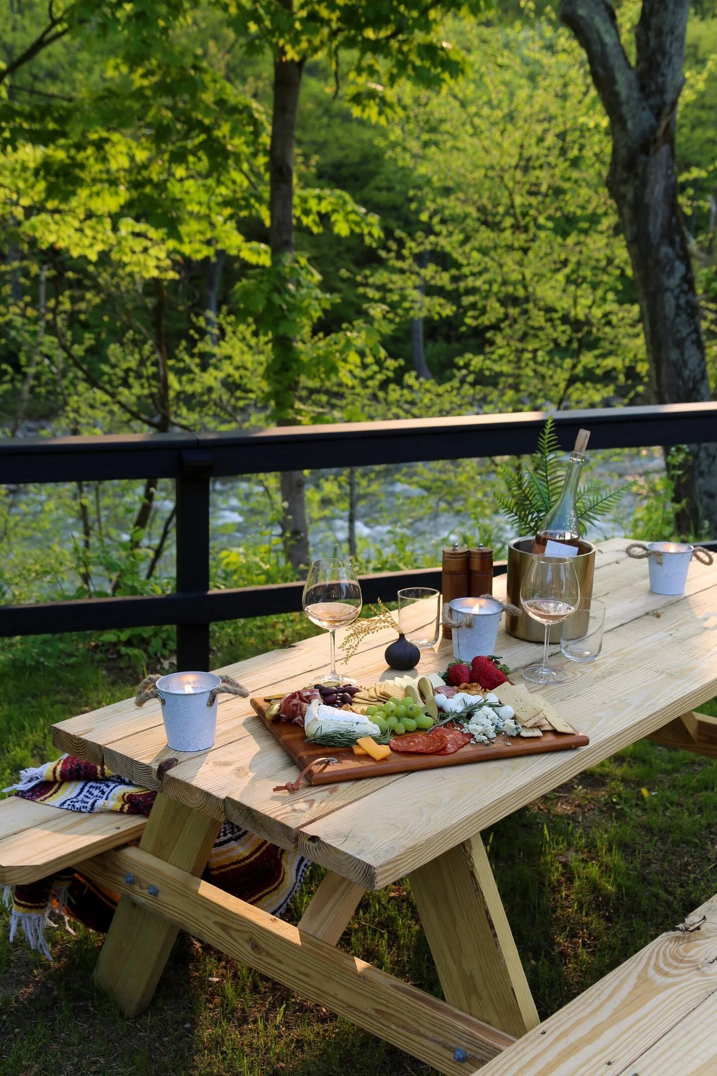 Picnic table set with a charcuterie board, wine glasses, candles, and a bottle, outdoors in a lush green forest.