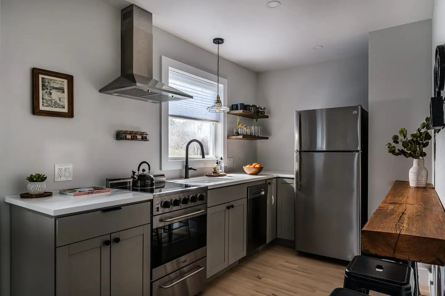 Modern kitchen with stainless steel refrigerator, stove, black kettle, white cabinets, wooden floating shelves, and a wooden side table with a plant in a vase.