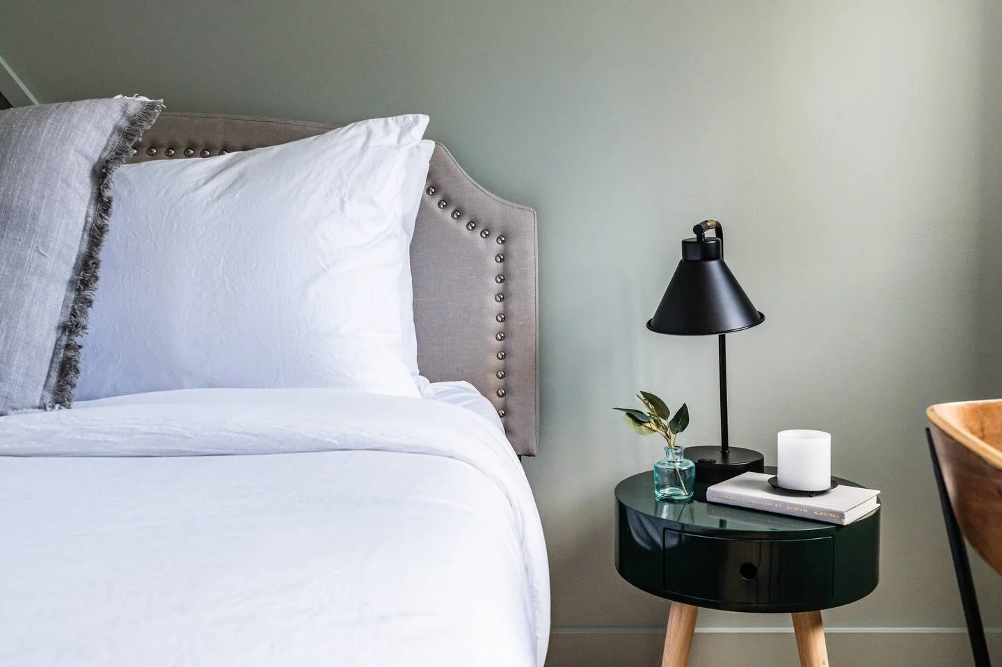 A neatly made bed with white bedding and decorative pillows next to a black nightstand holding a black table lamp, a small vase with greenery, and a candle.
