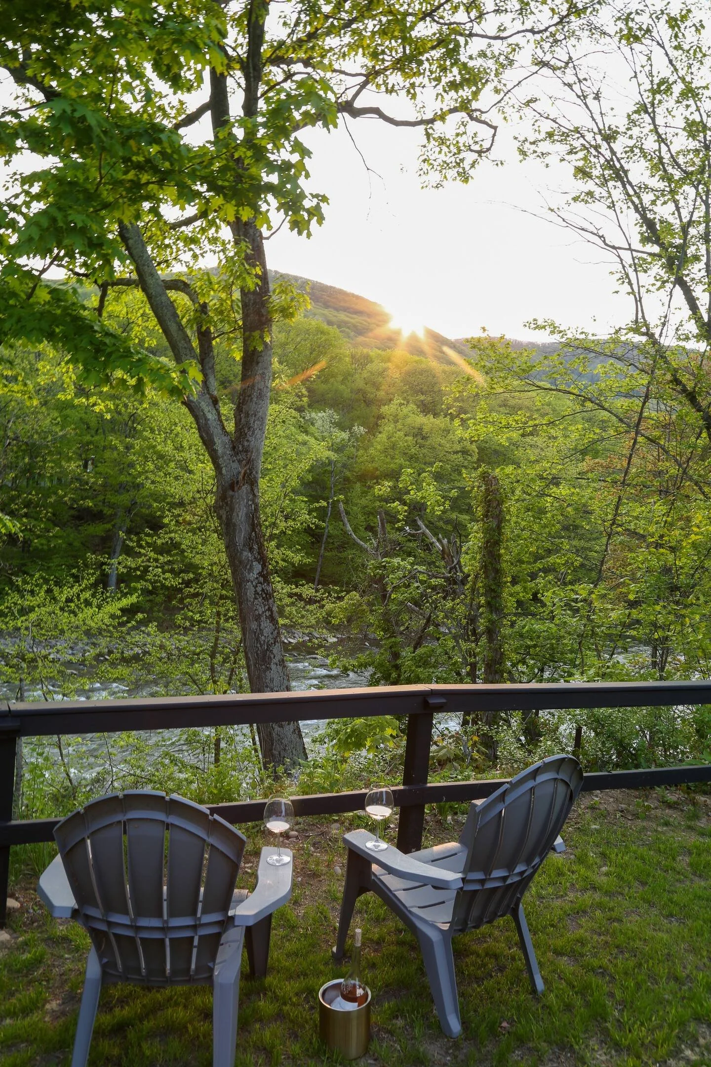 Two gray plastic chairs on a grassy porch with a small table holding two glasses of wine, overlooking a lush green forest with a river and hills in the distance at sunset.