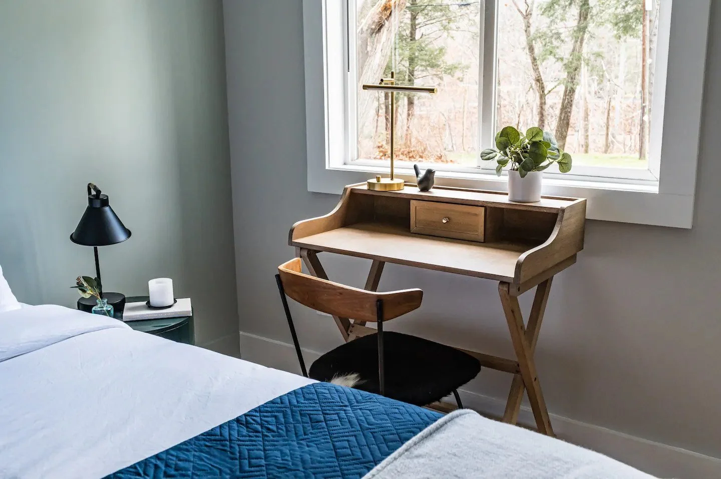 A cozy bedroom corner with a wooden desk, black chair, and window overlooking trees. The desk has a small drawer, a gold desk lamp, and a potted plant. A black table lamp and candles are on the bedside table. The bed has white sheets and a blue quilt.