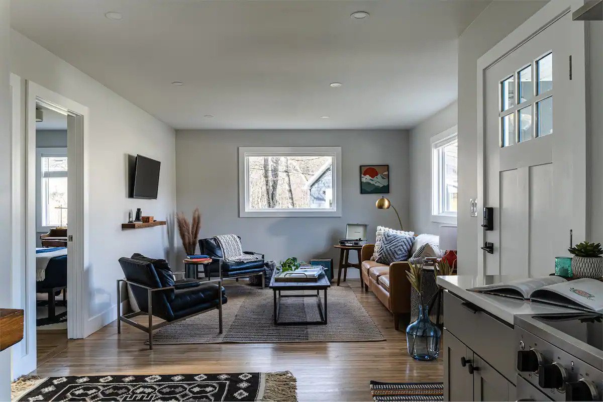 Living room with white walls, hardwood flooring, and natural light from windows. Contains a leather armchair and a sofa, side tables, wall-mounted TV, a small table with a lamp, artwork, and decorative plants.